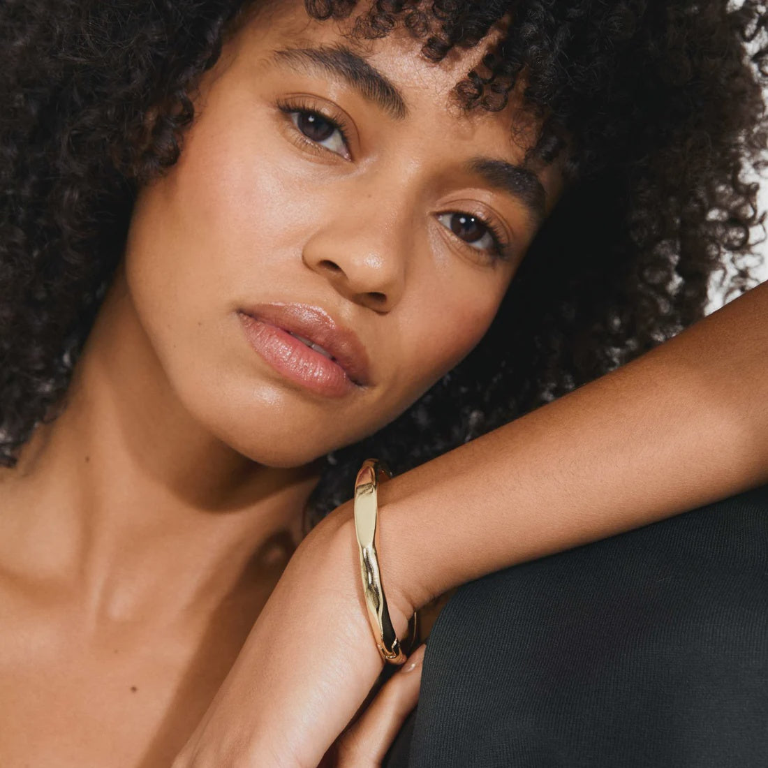 A close-up lifestyle shot of a model wearing a polished gold hinged bangle bracelet on her wrist, resting her arm on a dark surface.