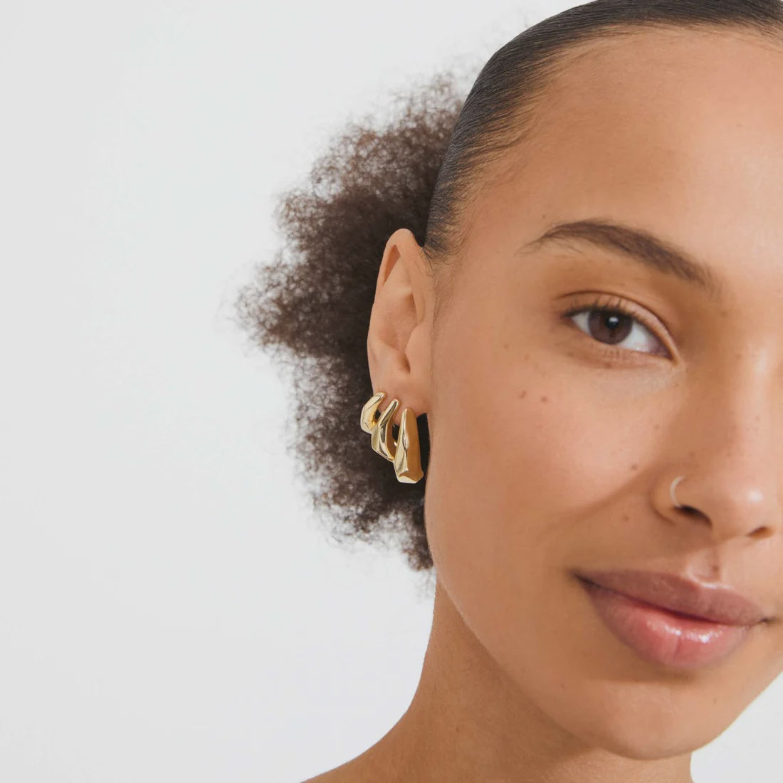 A close-up of a model wearing a pair of gold triple-hoop earrings against a clean white background.