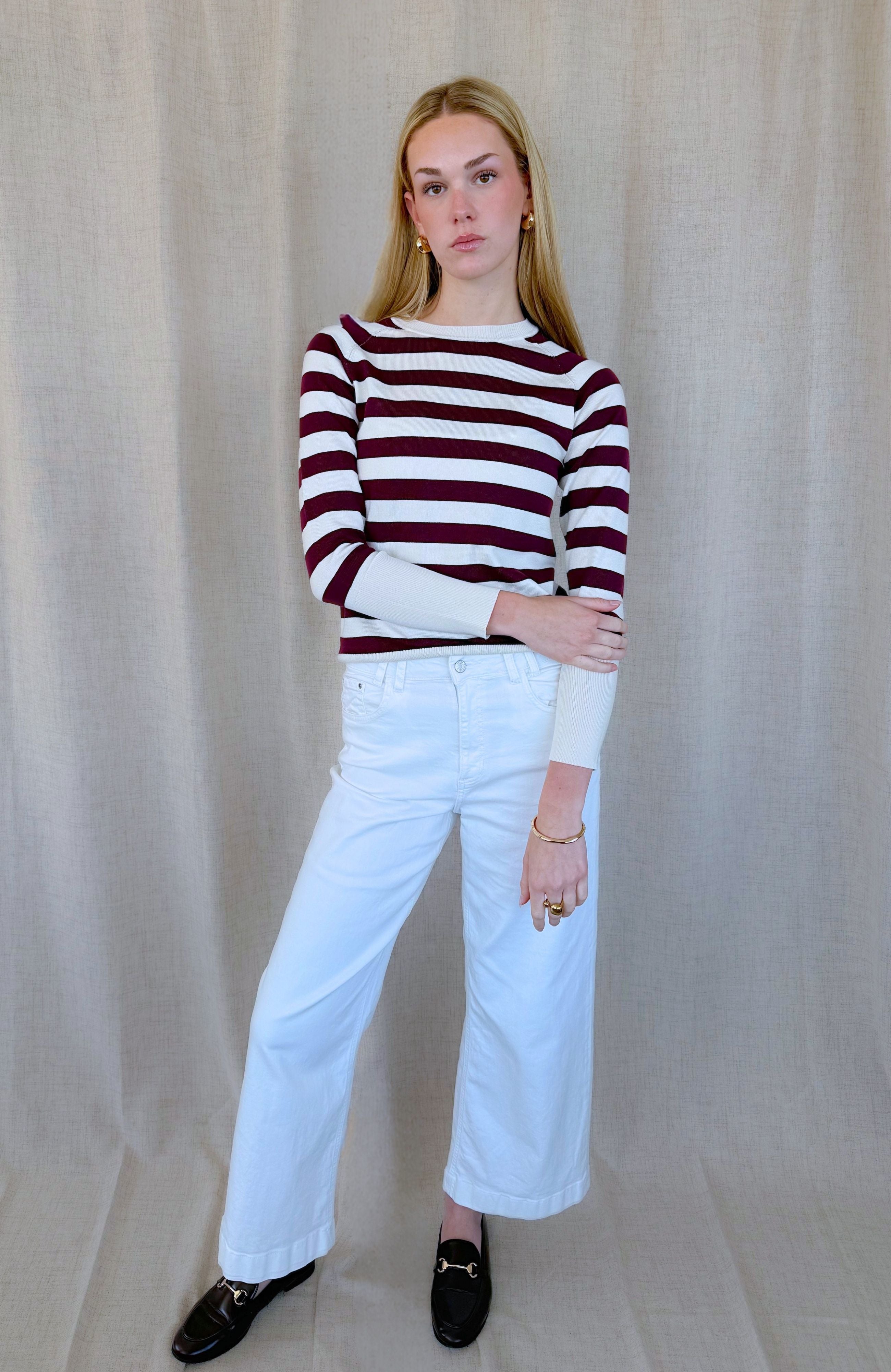 A woman modeling a long-sleeve knit top with horizontal burgundy and white stripes, paired with white wide-leg pants and black loafers, standing against a neutral beige fabric backdrop.