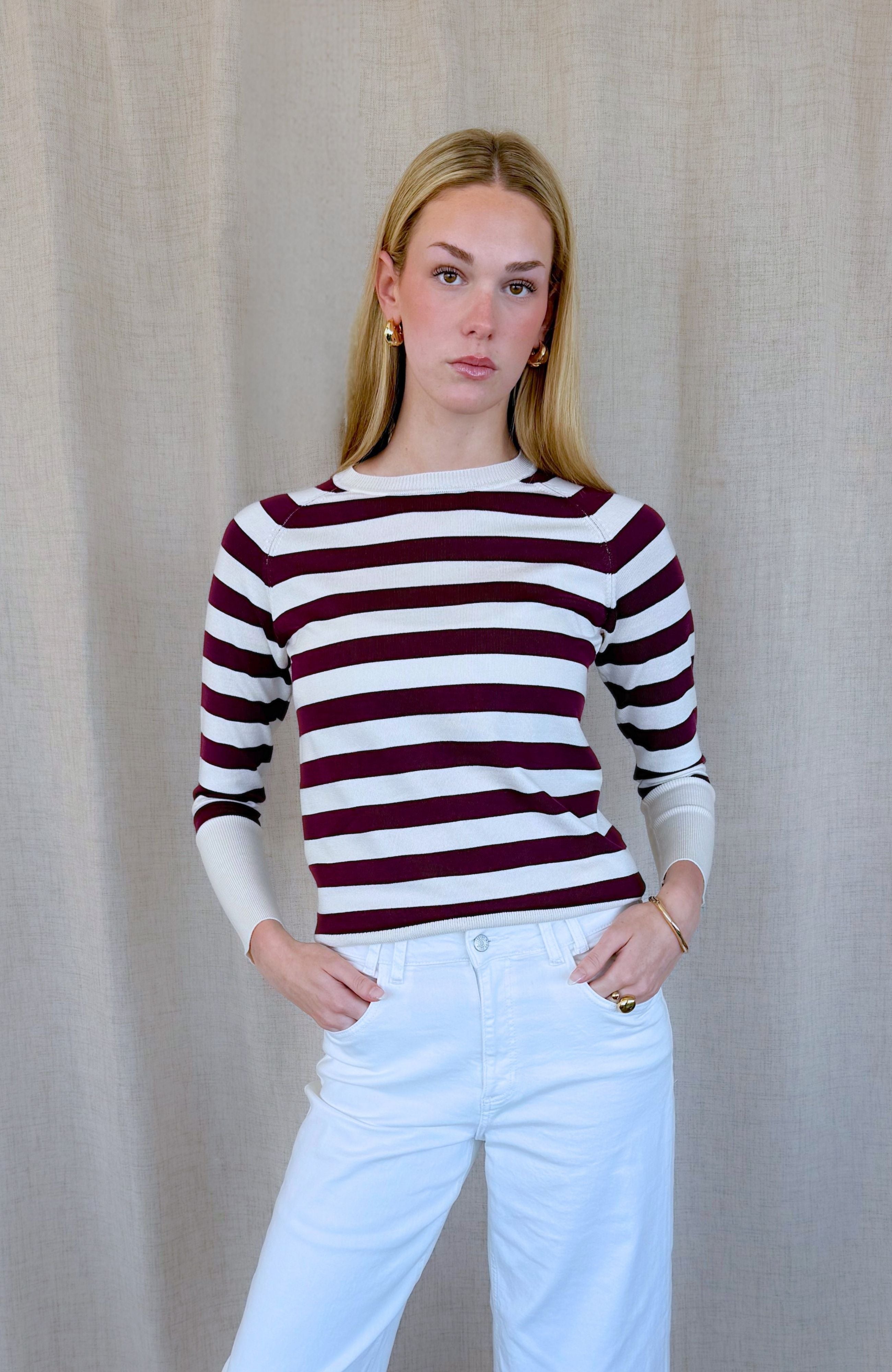 A woman modeling a long-sleeve knit top with horizontal burgundy and white stripes, paired with light blue jeans, standing against a neutral beige fabric backdrop.