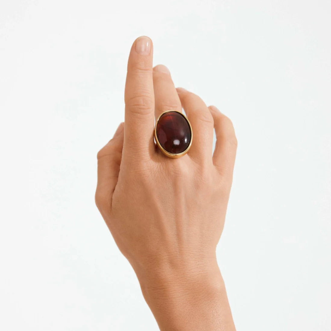 A gold ring with a large oval dark brown gemstone, worn on a person's finger against a clean white background.