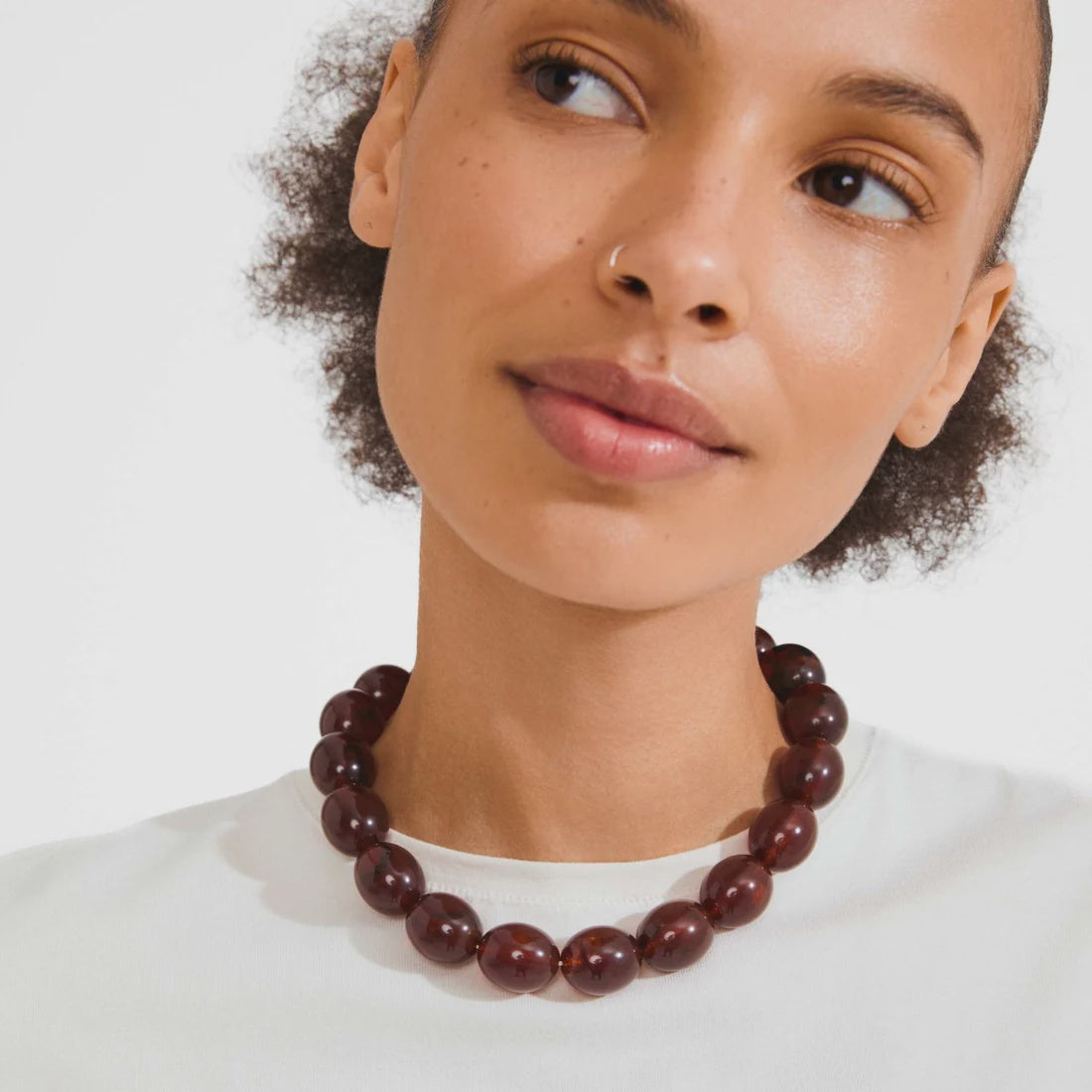 A close-up of a model wearing a dark reddish-brown beaded necklace with polished round beads, paired with a white crew-neck top against a light gray background.