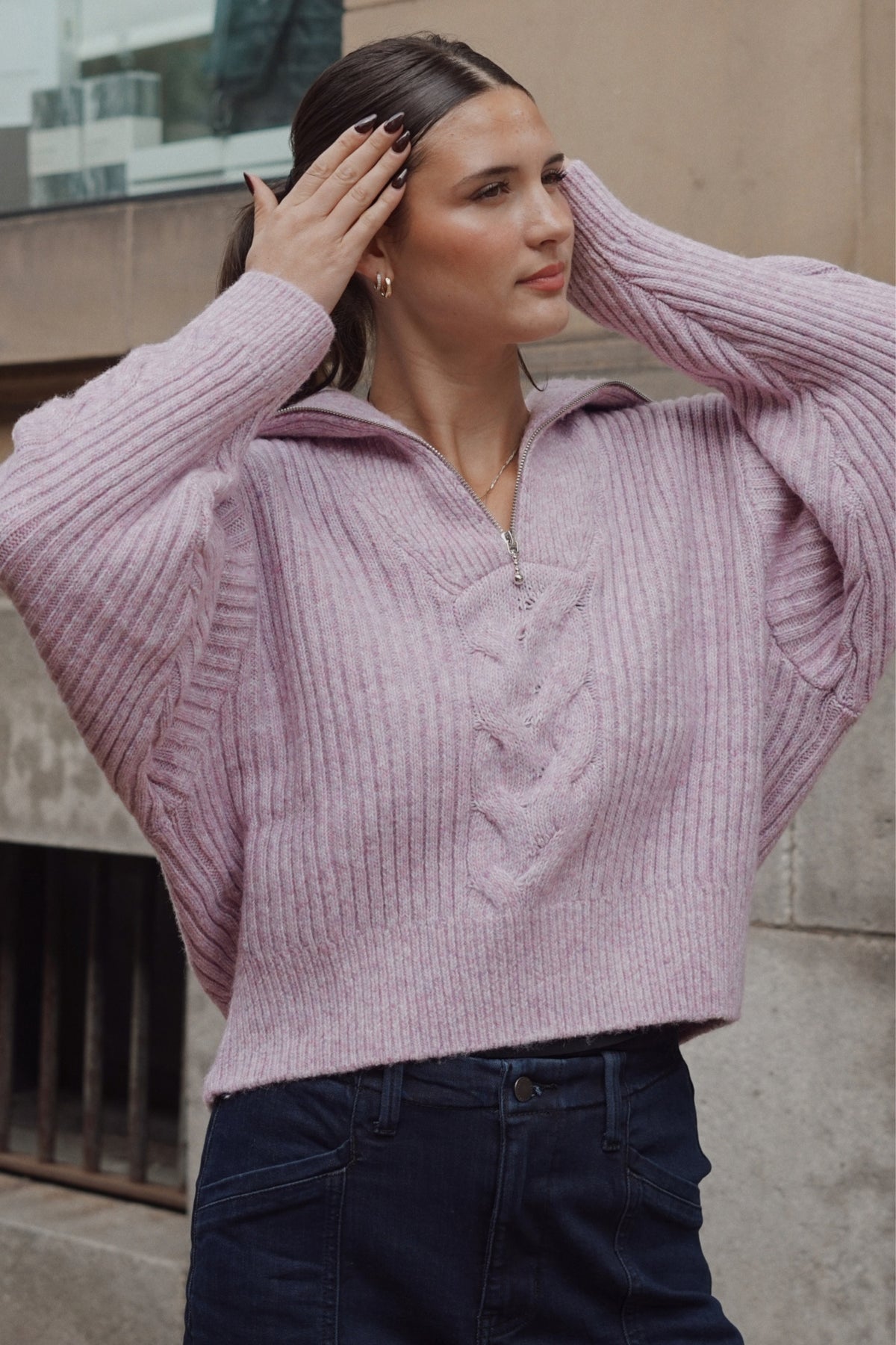A woman wearing a dusty pink ribbed knit half-zip sweater with a cable-knit front detail, paired with dark blue jeans, posing outdoors against a stone wall.