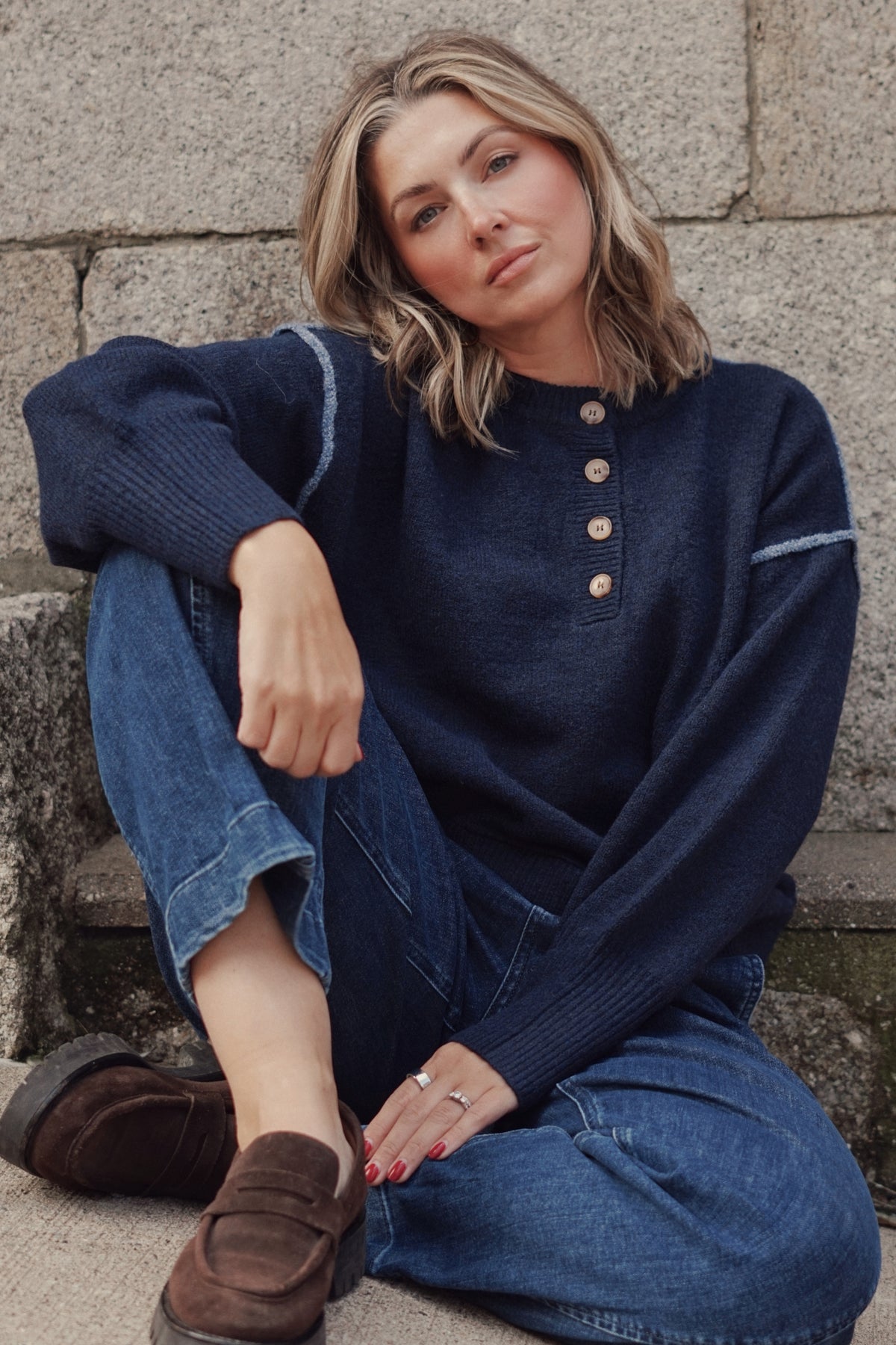 A woman wearing a navy blue knit sweater with a buttoned henley neckline, blue jeans, and brown suede loafers.