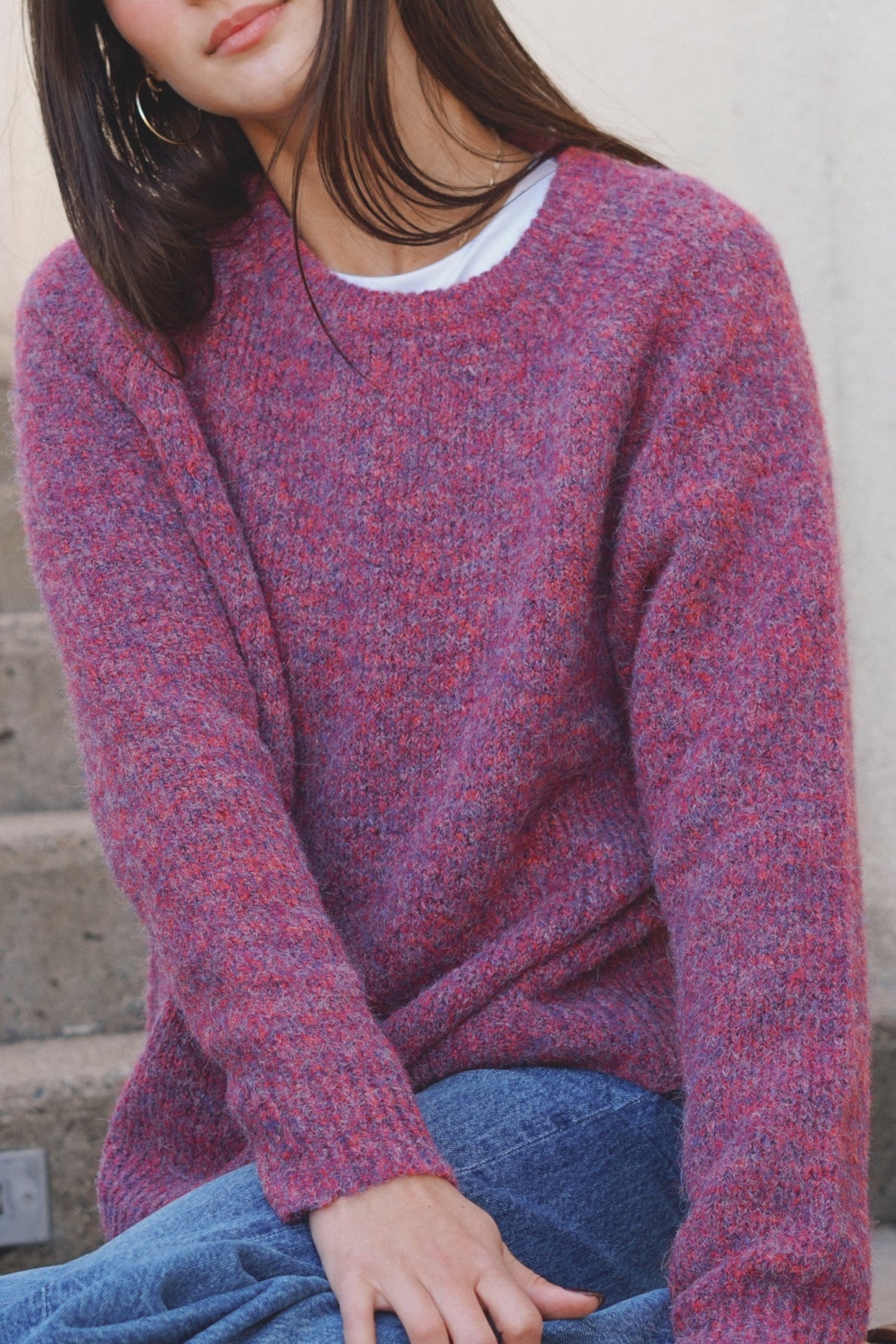 A woman wearing a fuzzy marled knit sweater in a dusty rose and red blend, paired with blue jeans, sitting on concrete steps.