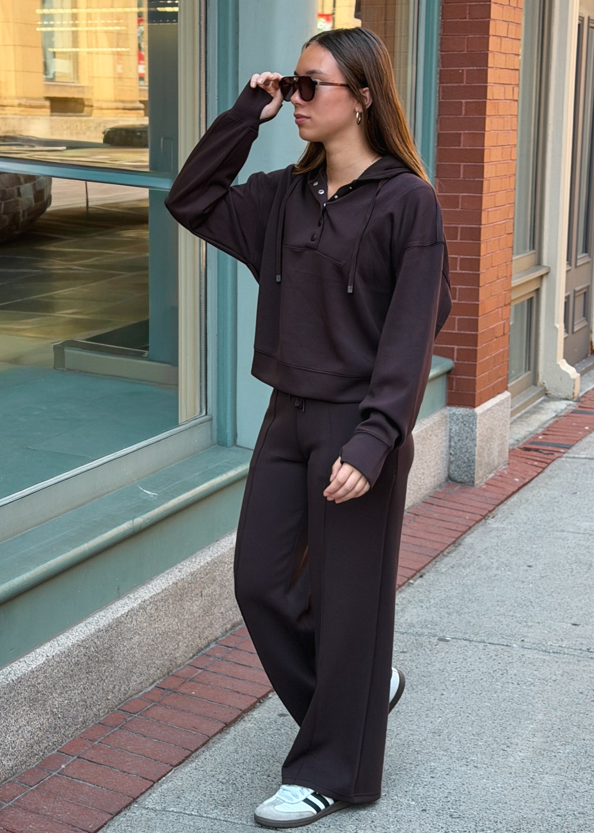 A woman wearing a matching dark brown lounge set consisting of a cropped hoodie and wide-leg sweatpants, walking on a city sidewalk next to a storefront.