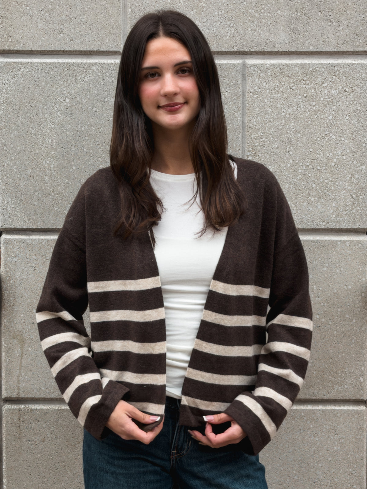 A woman wearing a brown and beige striped knit cardigan over a white top and blue jeans, standing against a gray stone wall.