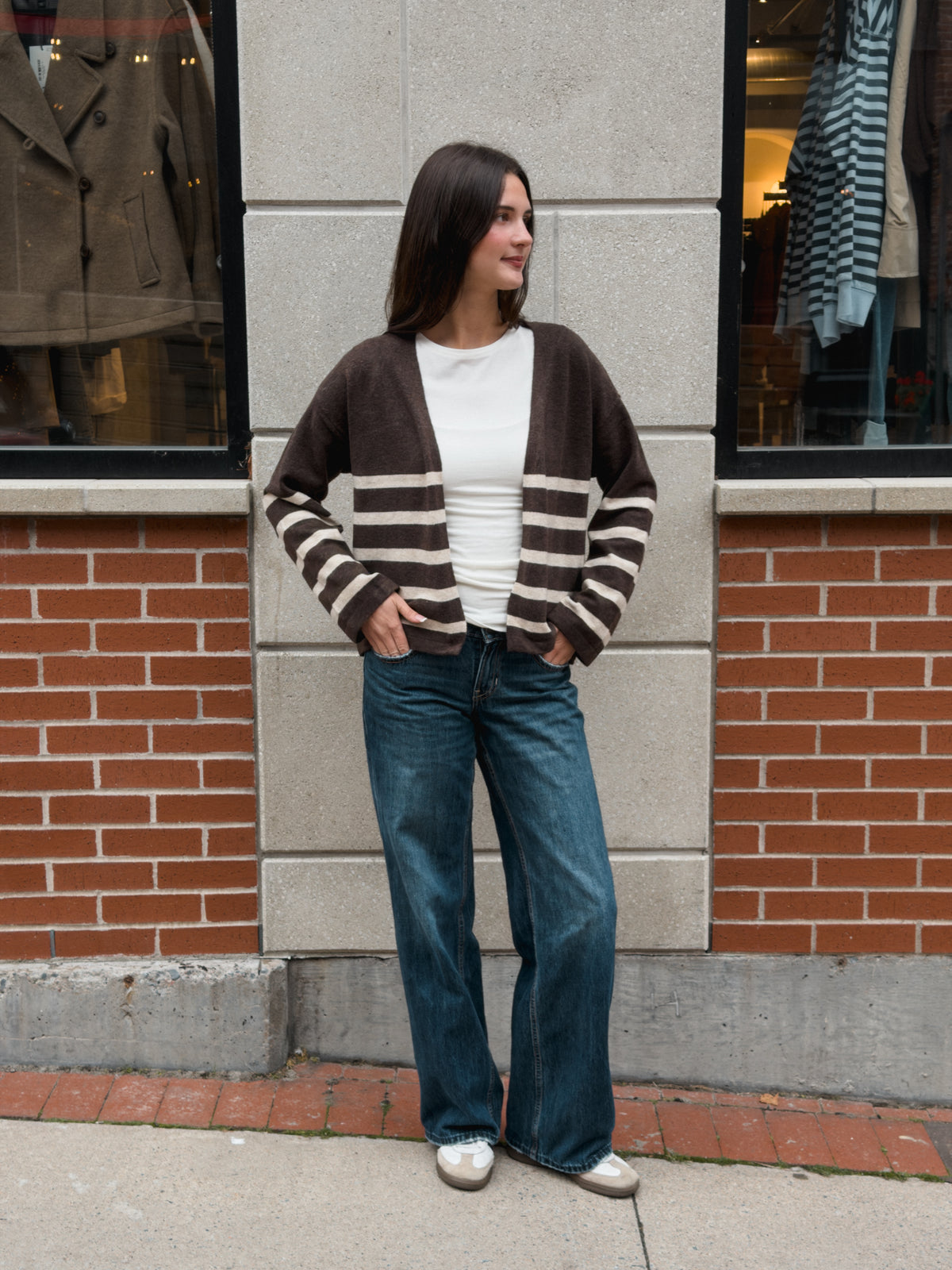 A woman stands on a sidewalk in front of a brick and stone building, wearing a brown and cream striped cardigan over a white top, blue wide-leg jeans, and white sneakers.