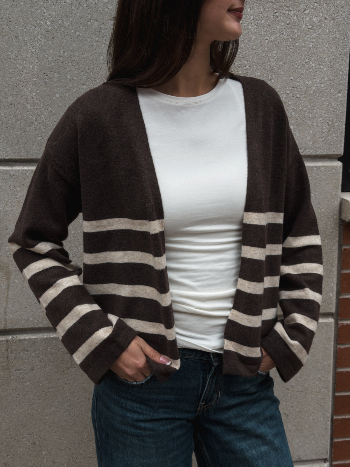 A woman wearing a brown open-front cardigan with beige horizontal stripes on the sleeves and hem, layered over a white t-shirt and blue jeans, standing against a gray stone wall.