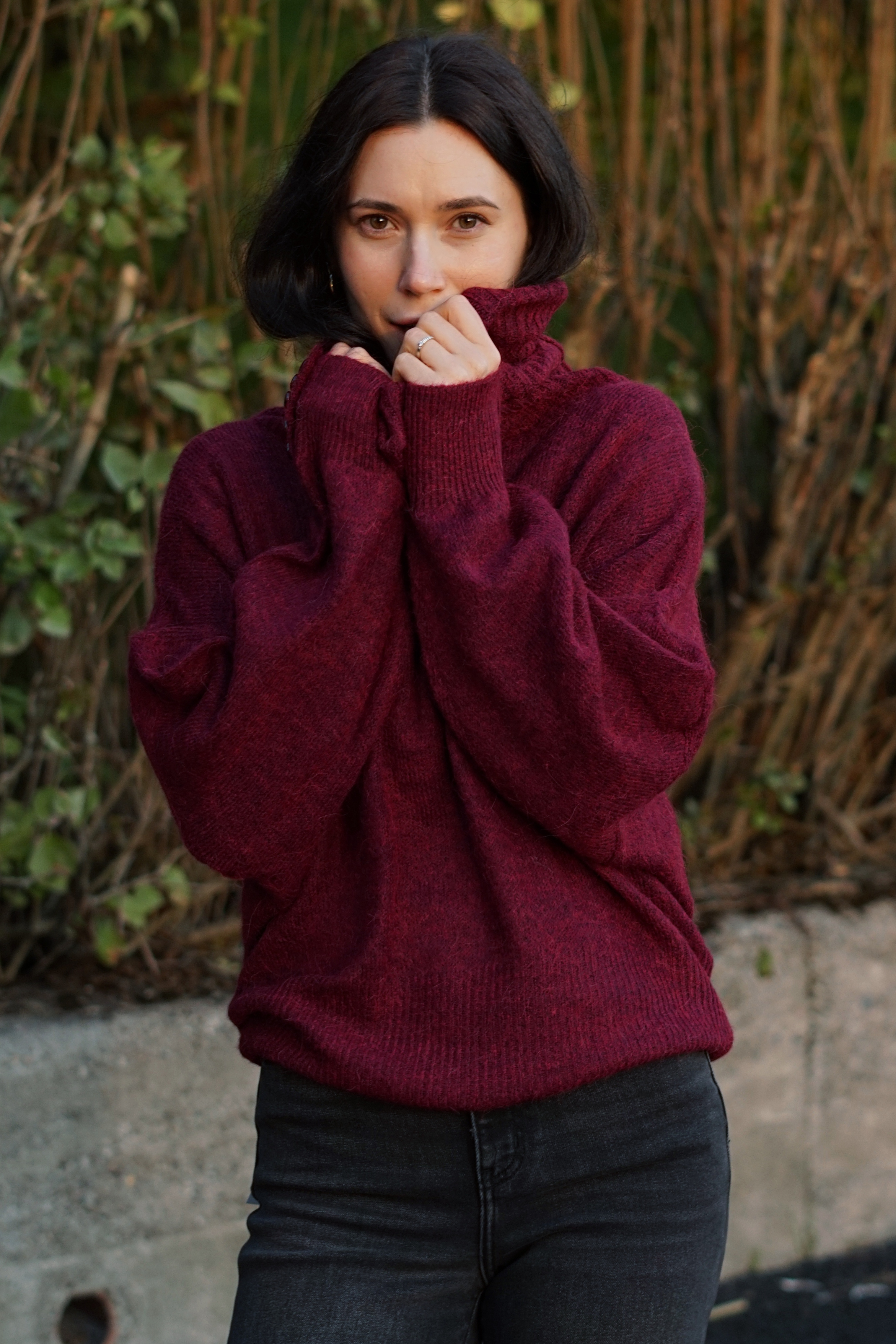 A woman wearing a chunky, oversized burgundy turtleneck sweater and dark gray jeans, standing outdoors in front of a bamboo fence.