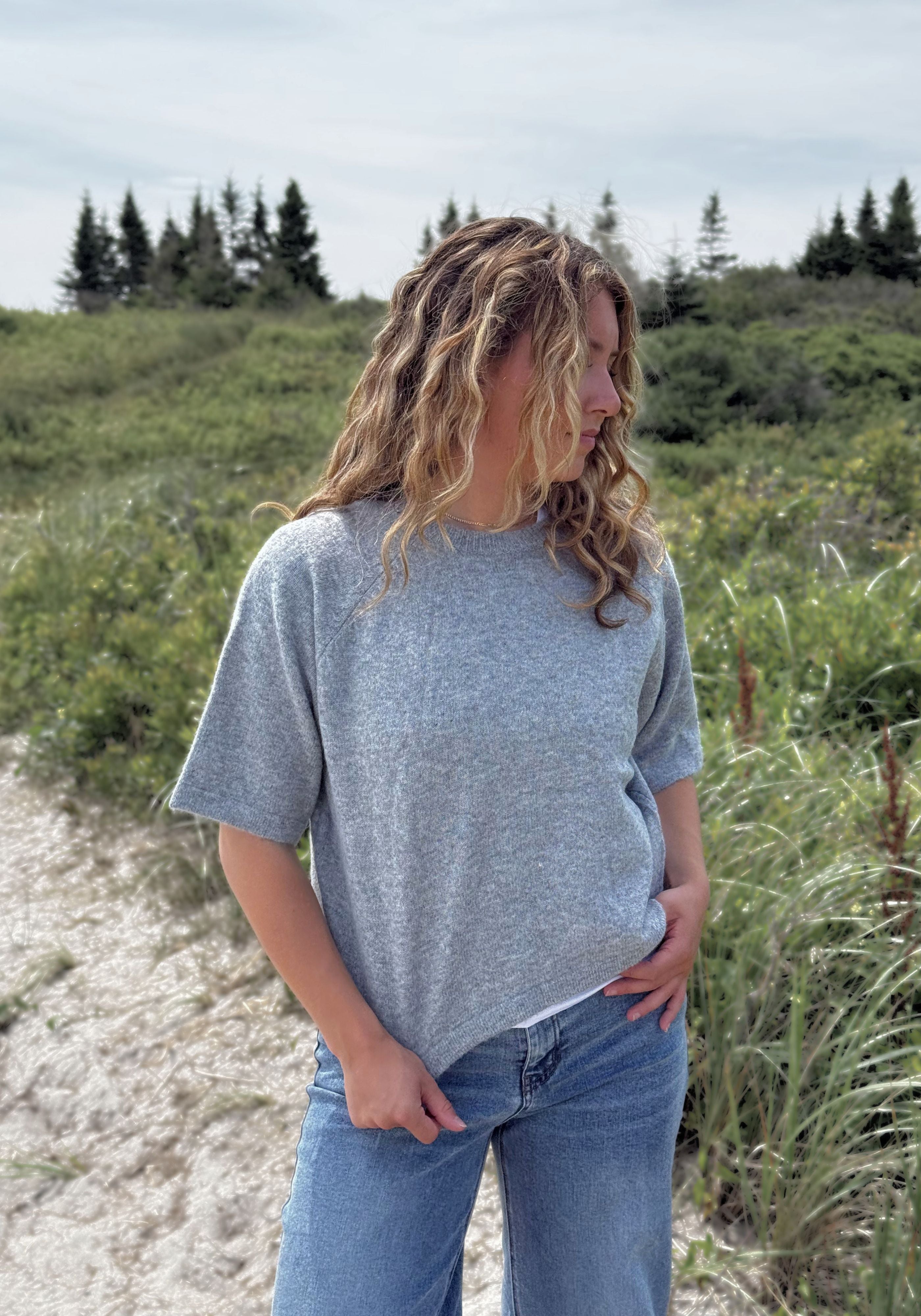 A woman wearing a heather gray short-sleeve t-shirt and blue jeans standing outdoors on a sandy path.