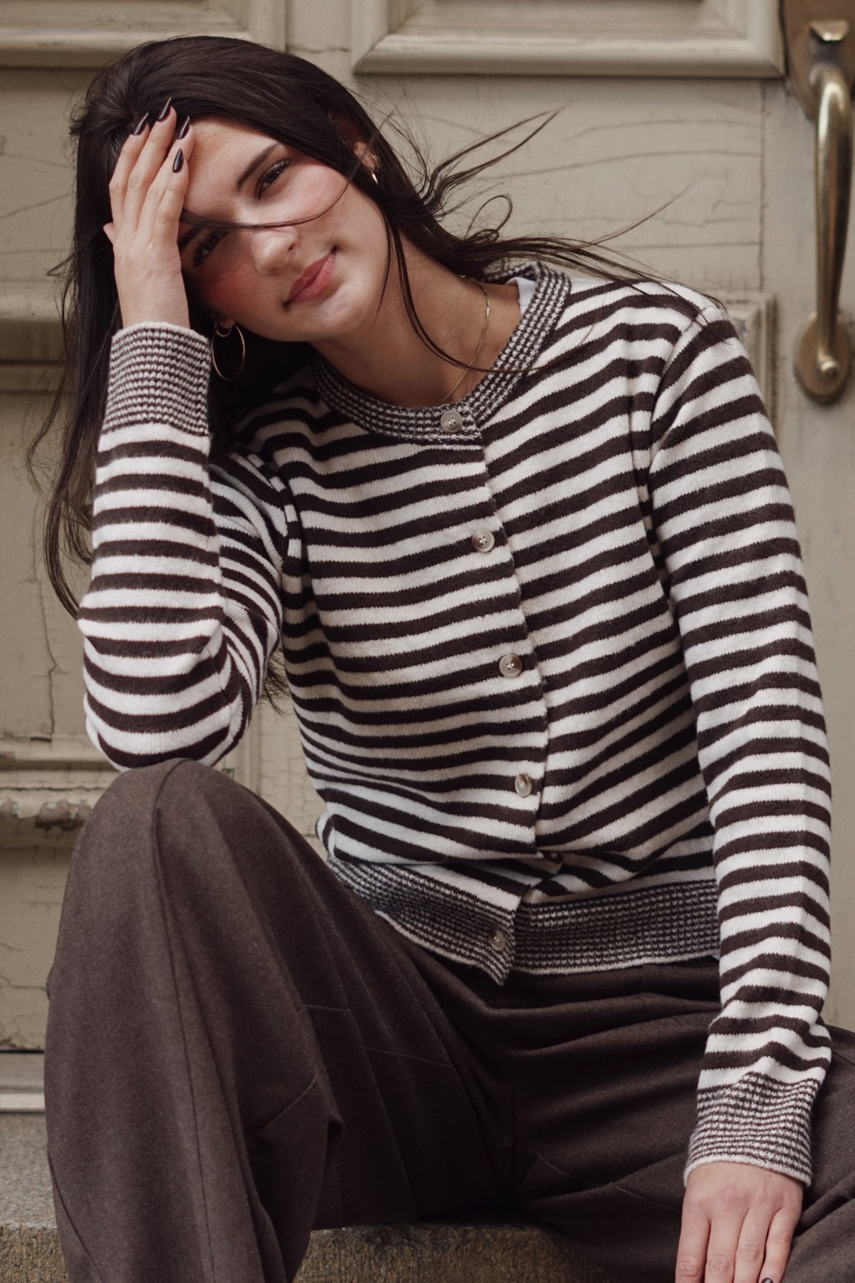 A woman wearing a brown and cream striped knit cardigan sweater and dark brown pants, sitting on stone steps.