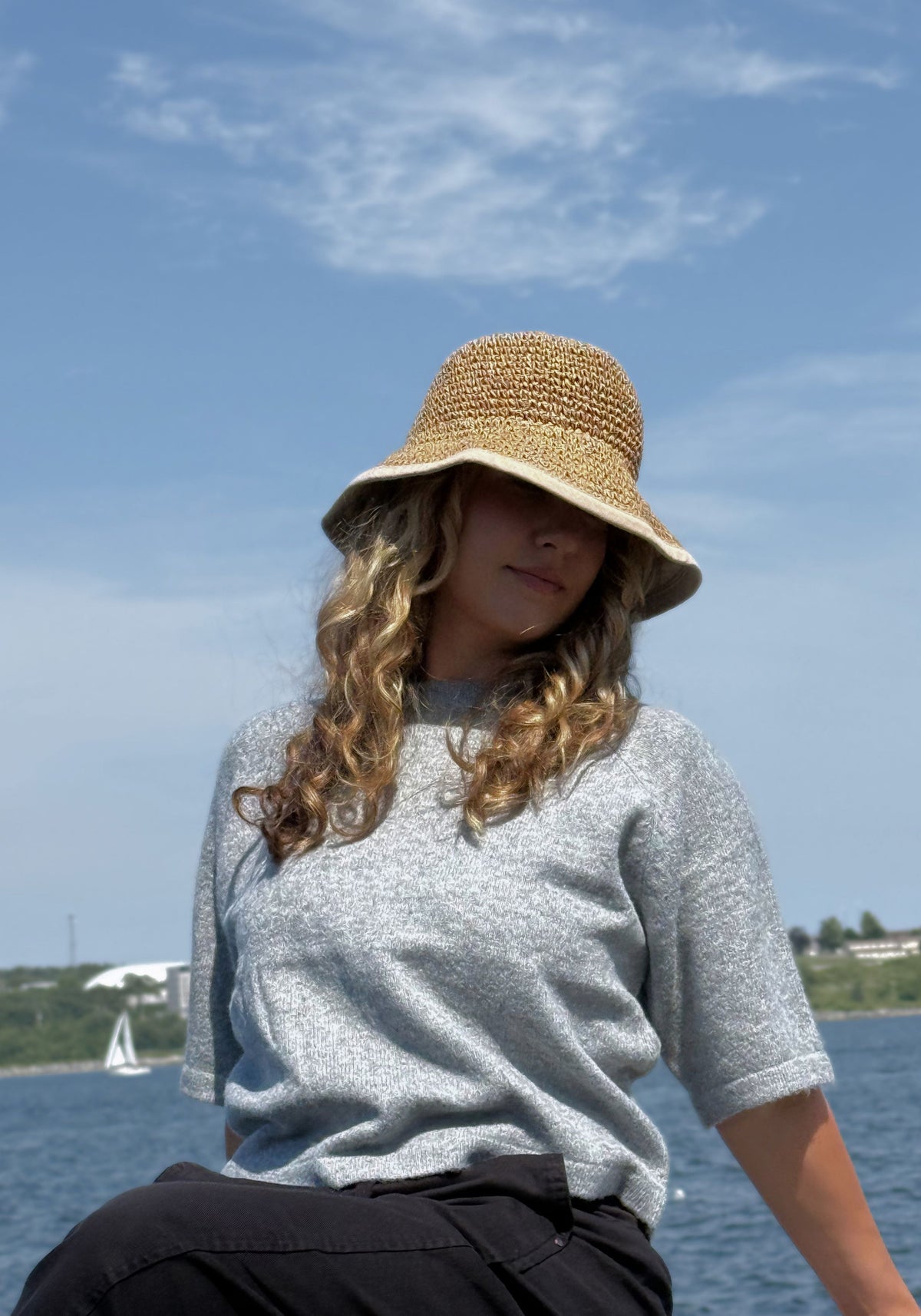 A woman wearing a wide-brimmed straw sun hat, a light gray knit top, and black pants, sitting outdoors.