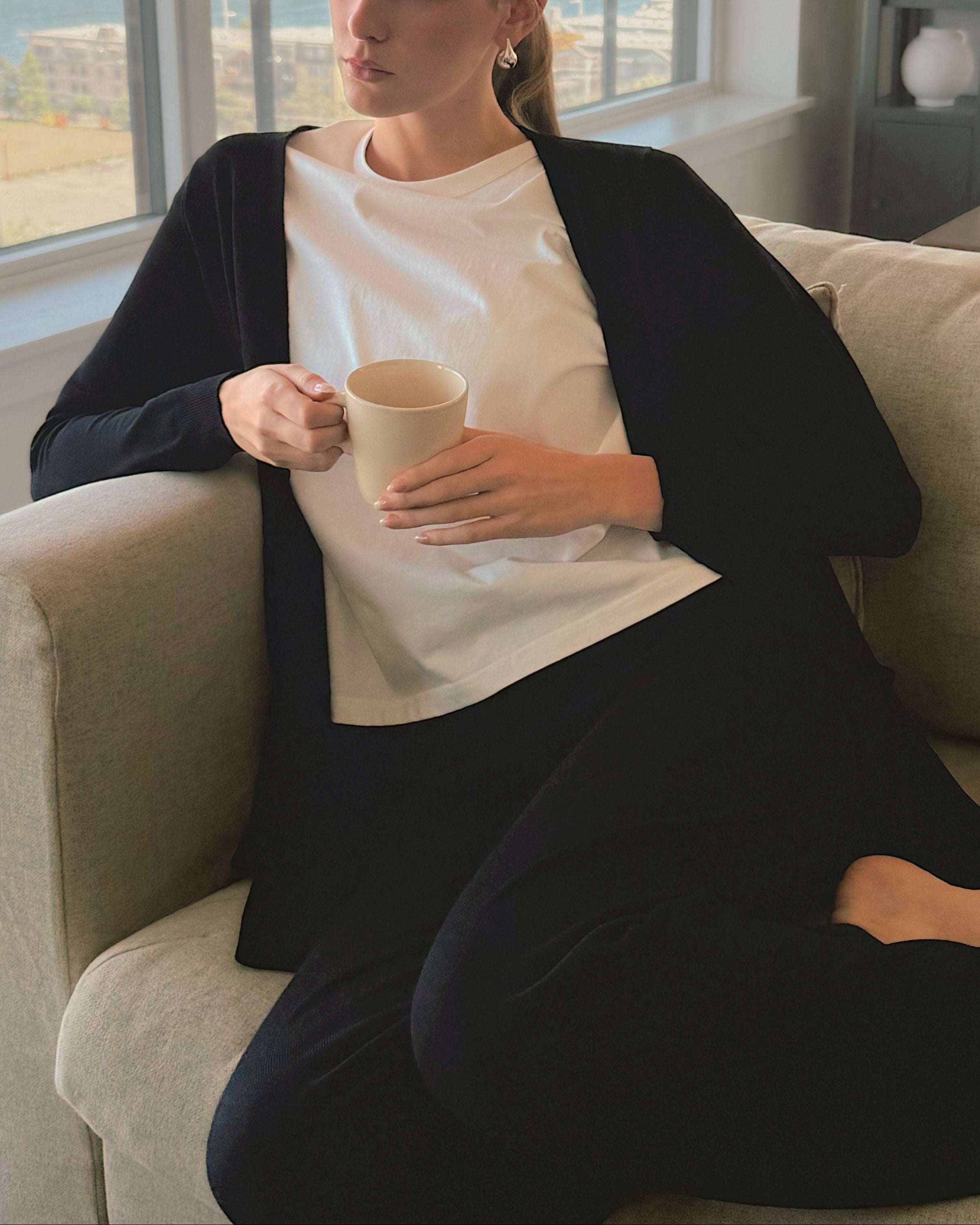 A woman sits on a light beige sofa in a bright room, wearing a white crewneck t-shirt under a black cardigan and black pants, holding a beige ceramic mug.