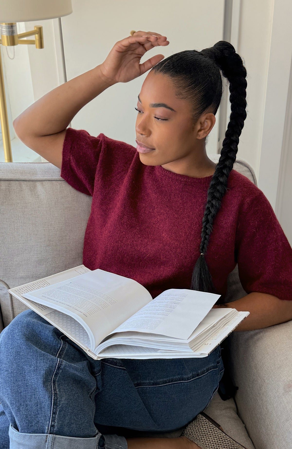 A woman with a long black braided ponytail sits on a light gray couch, wearing a heathered burgundy short-sleeve top and blue jeans, holding an open book.