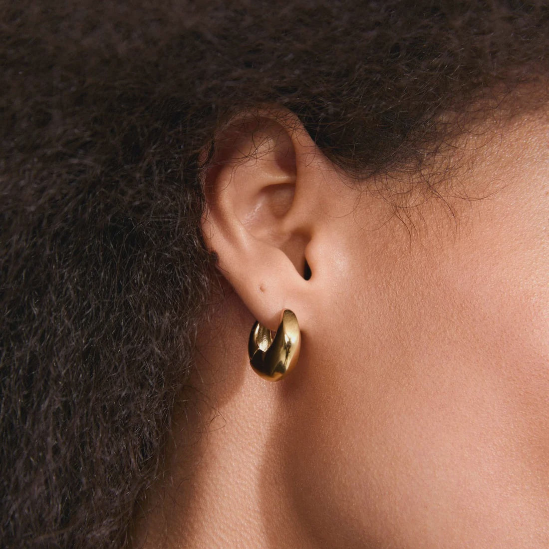 A close-up of a person's ear wearing a small polished gold hoop earring.
