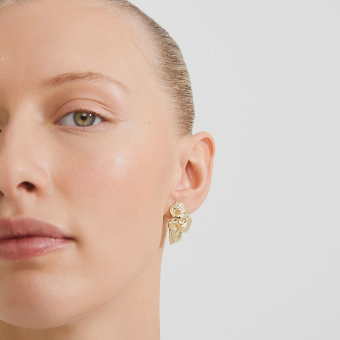 A close-up of a model's ear wearing a gold-toned, organic-shaped stud earring with a small clear stone accent, set against a plain light gray background.