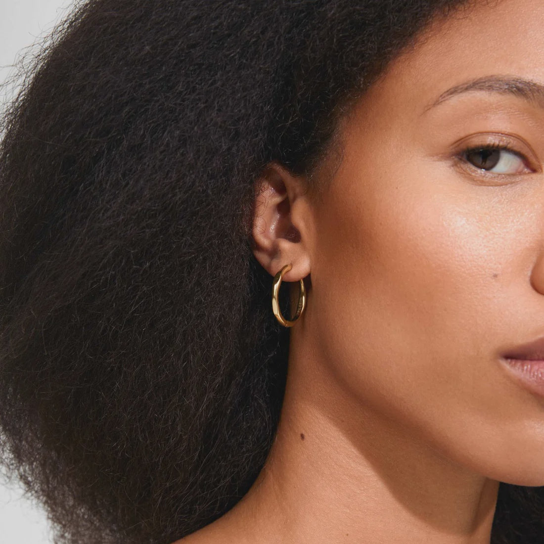 A close-up of a woman with dark curly hair wearing a thin gold hoop earring.