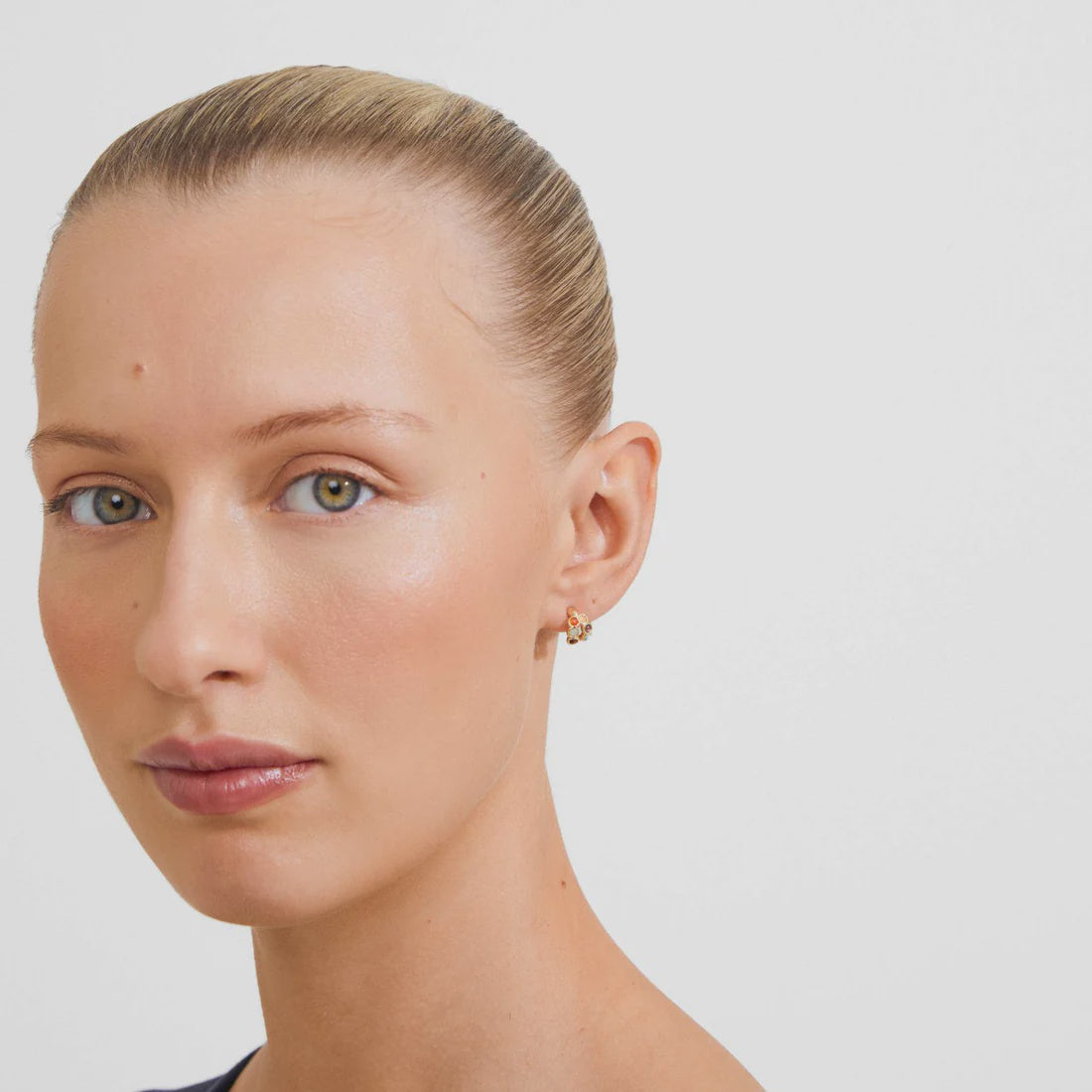 A close-up portrait of a woman with her hair pulled back, wearing a small gold hoop earring on her earlobe, against a plain light gray background.