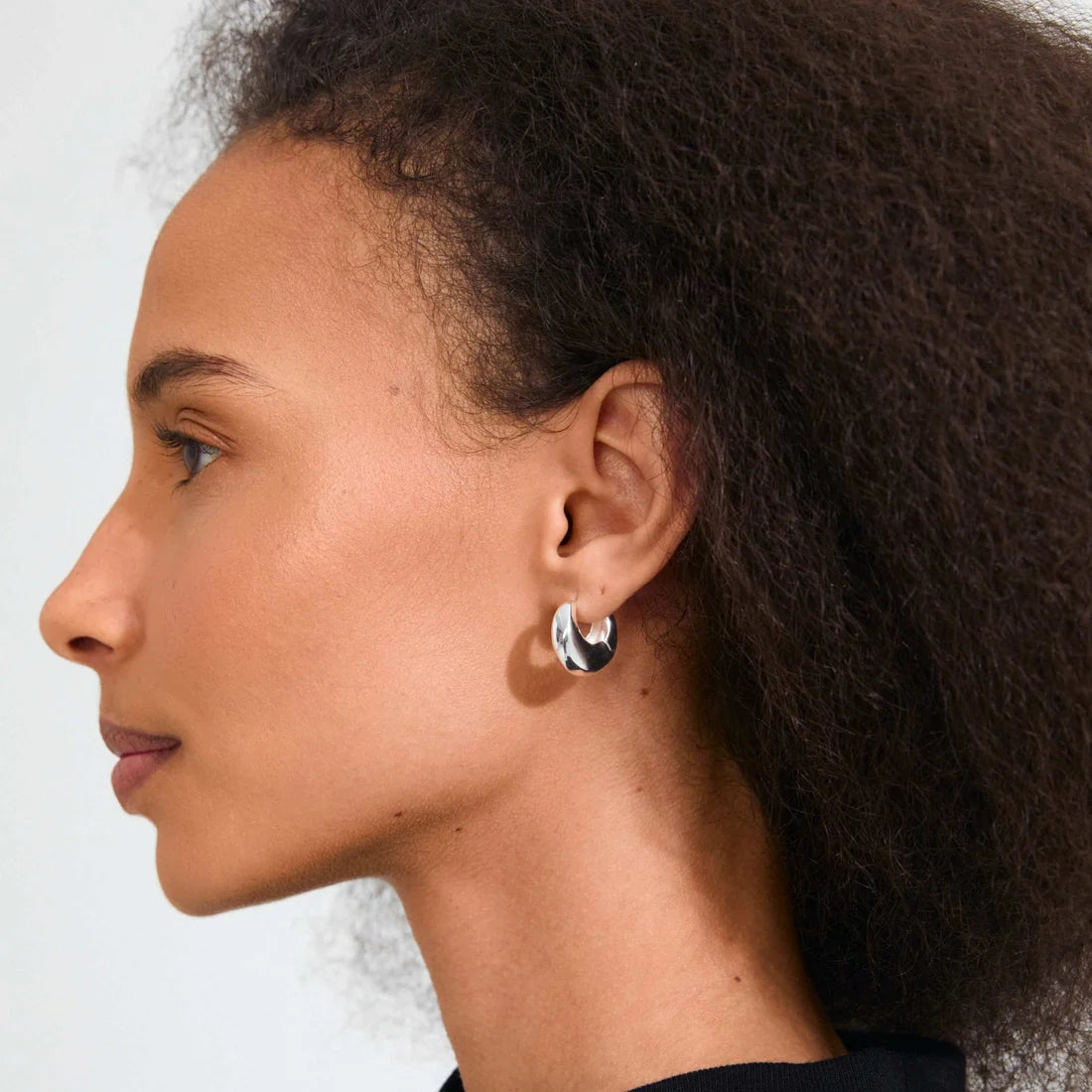 A close-up side profile of a woman with dark curly hair wearing a small silver hoop earring with a black and white patterned center, against a light gray background.
