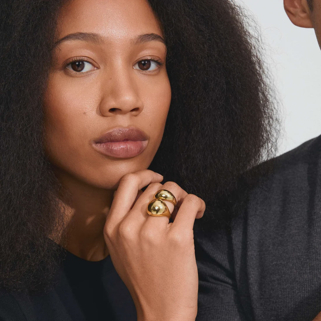 A close-up lifestyle shot of a model wearing a chunky gold ring with two stacked, rounded gold discs on her finger, set against a dark gray knit sweater and a light background.