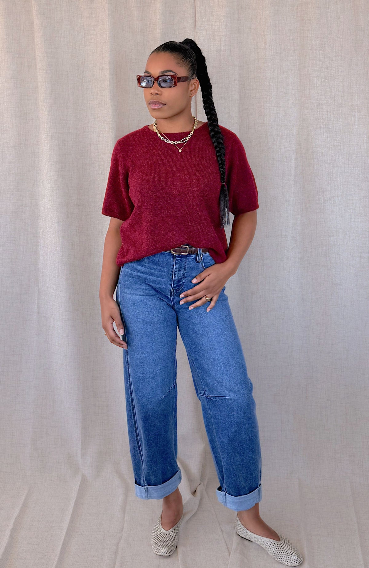 A woman modeling a loose-fitting maroon short-sleeve knit top, blue wide-leg jeans, and silver flat shoes, standing against a neutral draped fabric backdrop.
