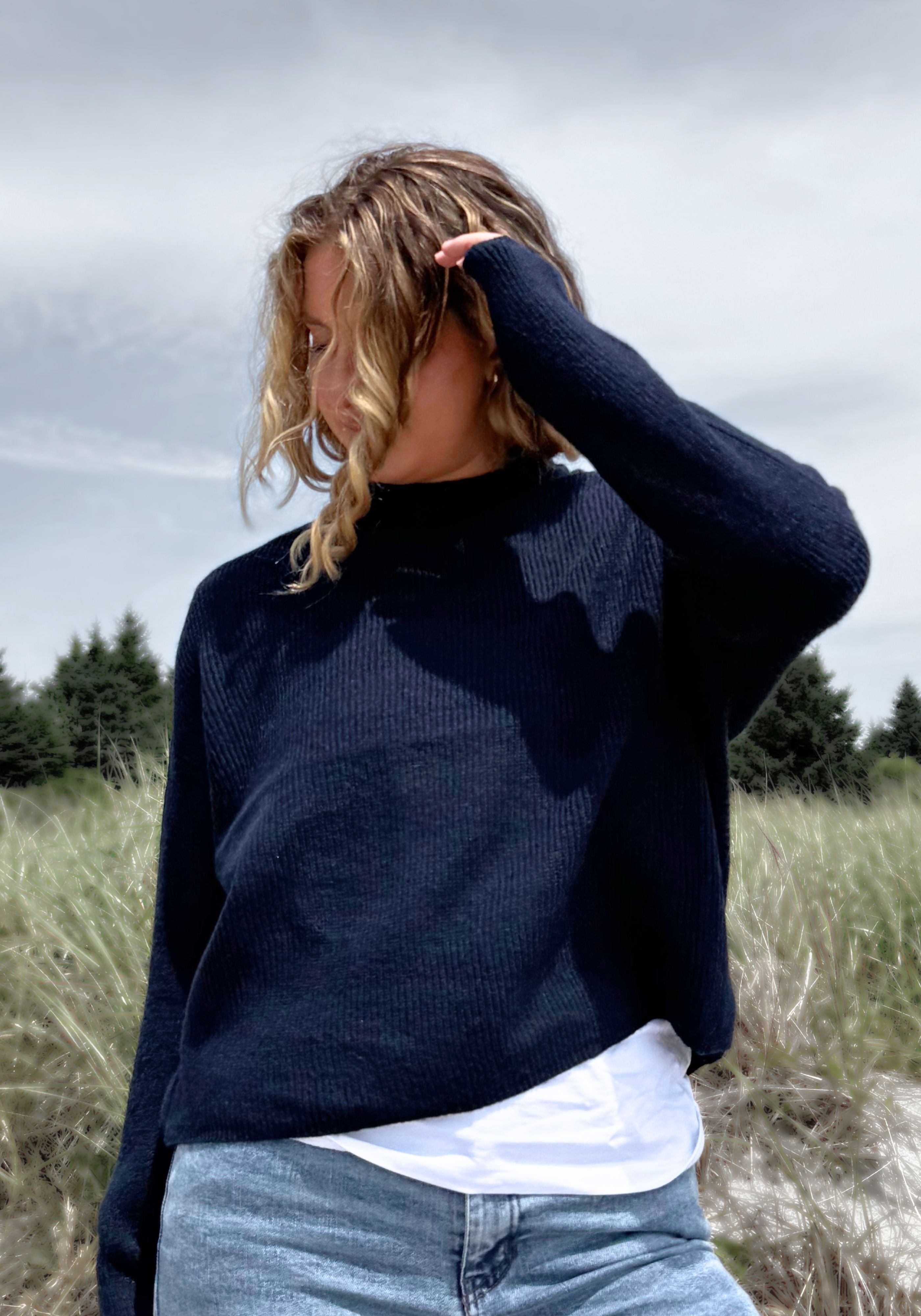 A woman wearing a dark navy ribbed knit mock neck sweater, a white t-shirt, and light blue jeans, standing in an outdoor grassy field under a cloudy sky.