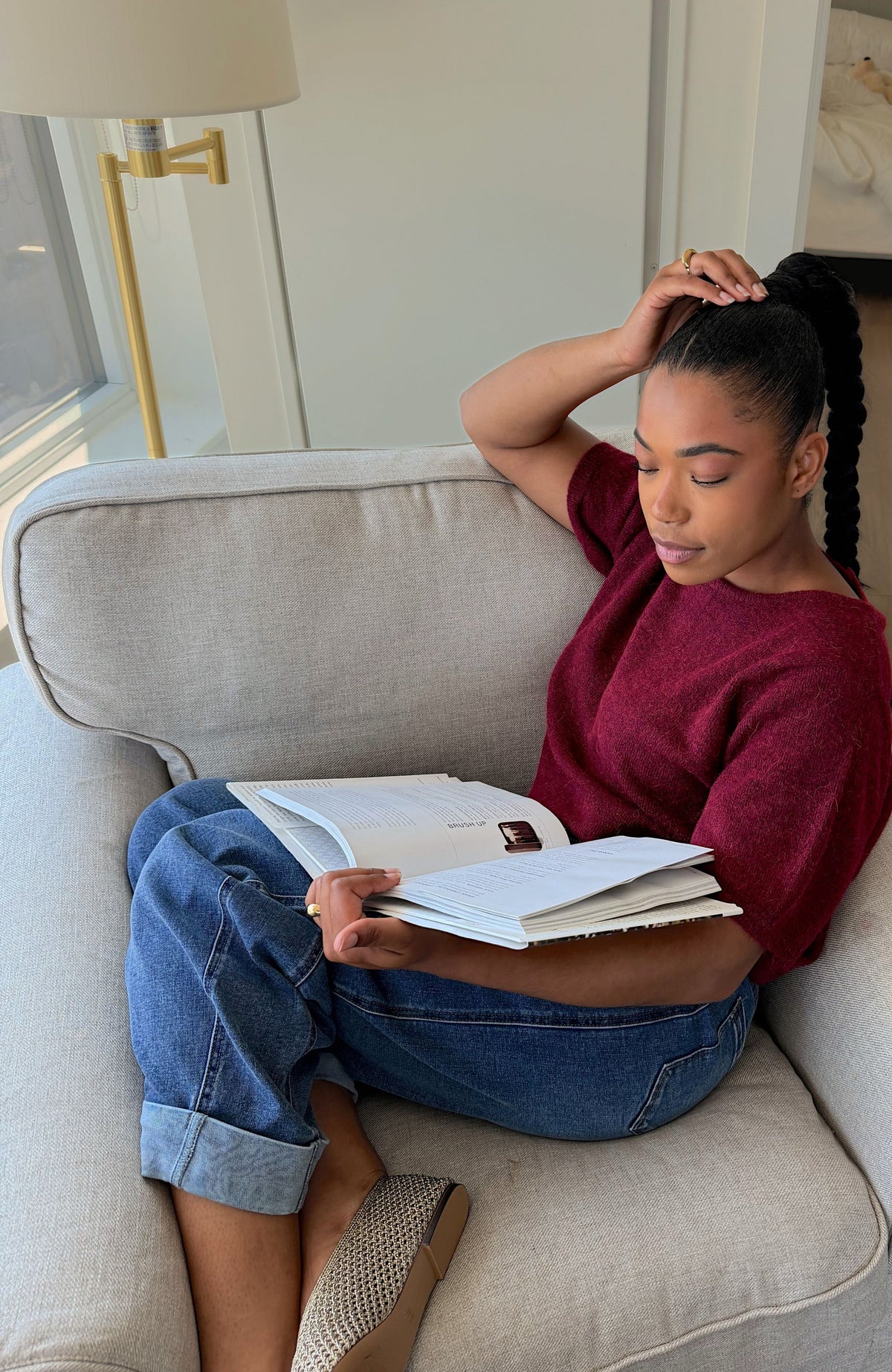 A woman sits on a light beige armchair in a bright room, holding an open book. She is wearing a maroon short-sleeve top, blue jeans, and textured tan shoes.