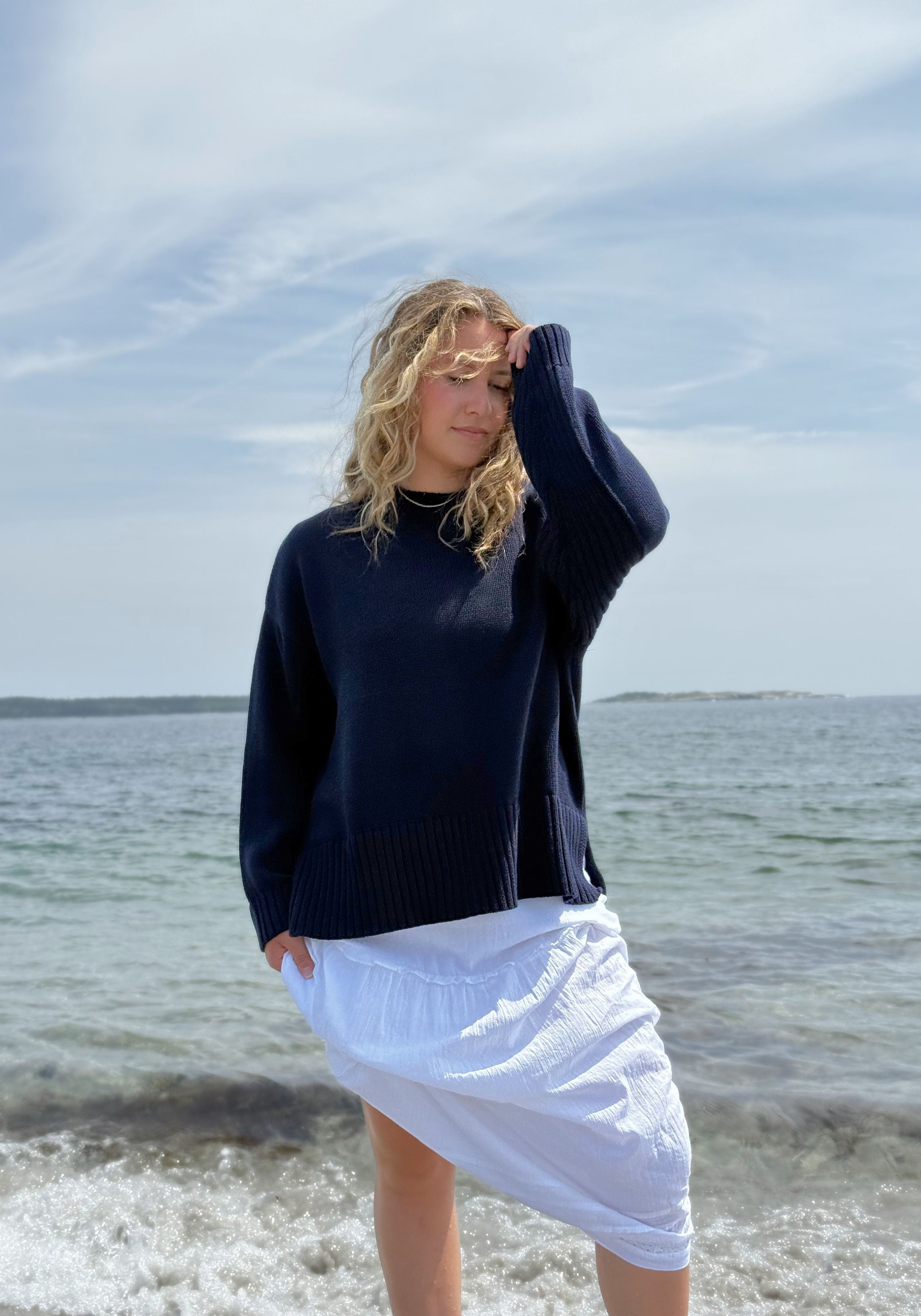 A woman standing on a beach wearing a dark navy blue knit sweater and a white skirt.