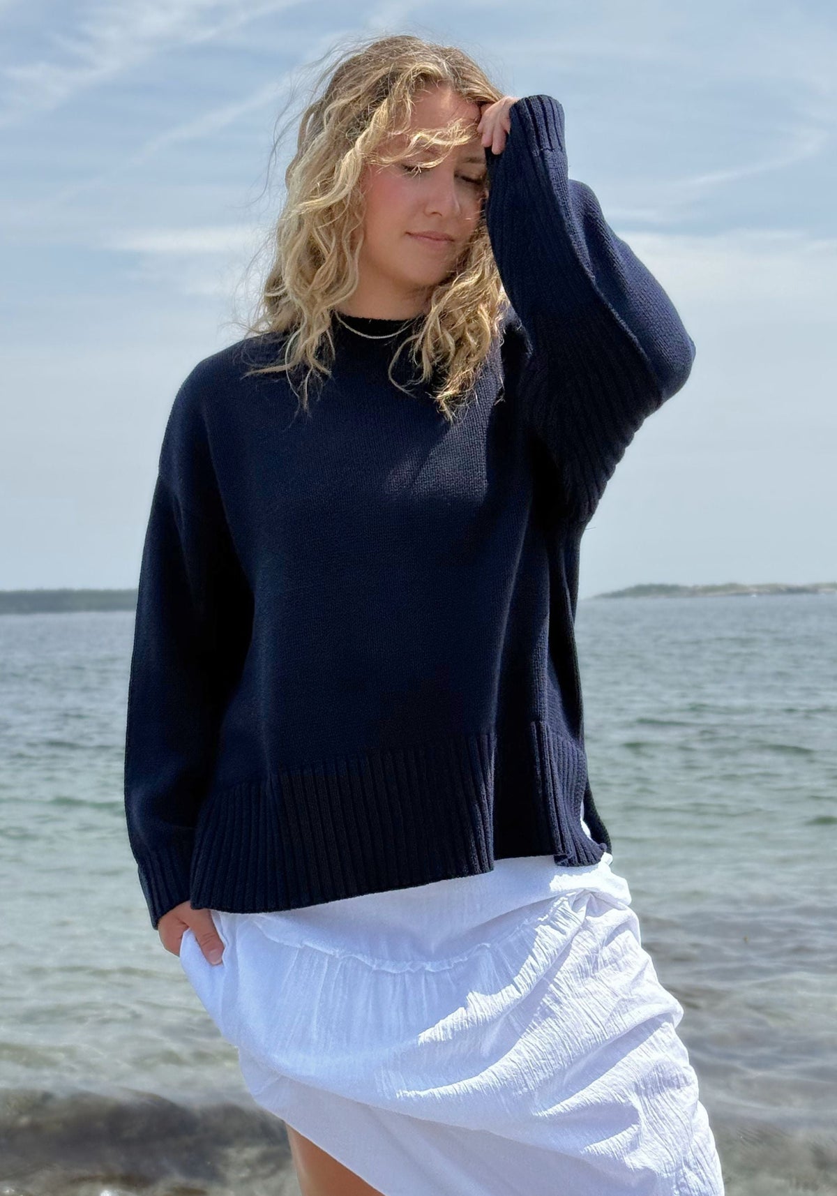 A woman standing on a beach wearing a dark navy blue knit sweater and a white skirt.