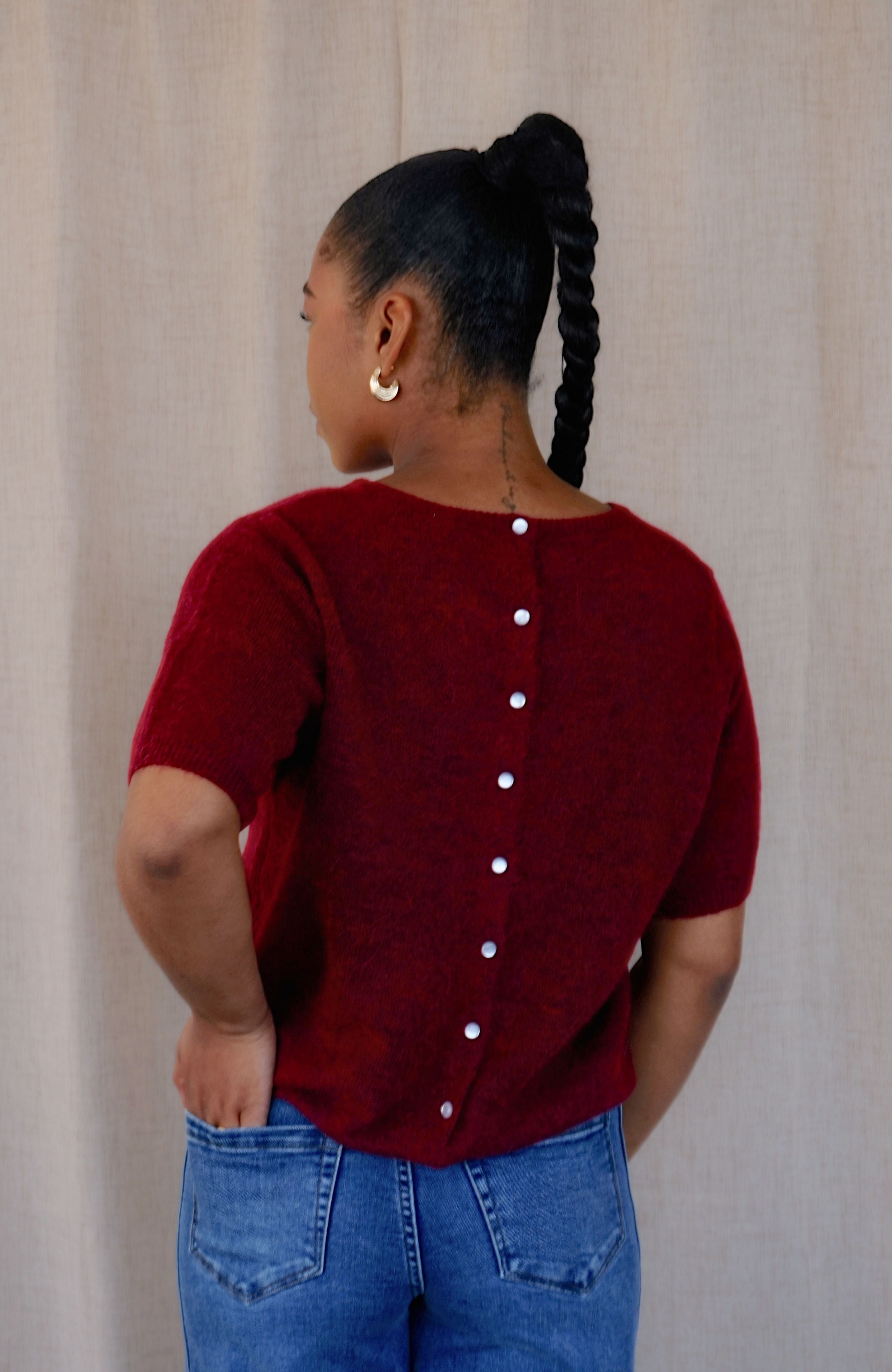 A woman shown from the back wearing a short-sleeve burgundy knit top with a vertical row of white buttons down the back, paired with blue jeans, against a neutral beige fabric backdrop.
