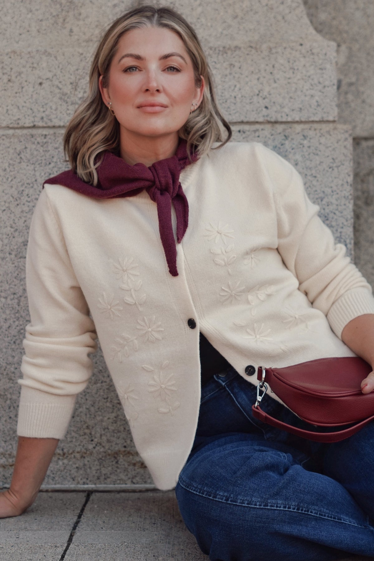 A woman wearing a cream-colored knit cardigan with tonal floral embroidery, a dark burgundy neck scarf, blue jeans, and carrying a matching burgundy crossbody bag.