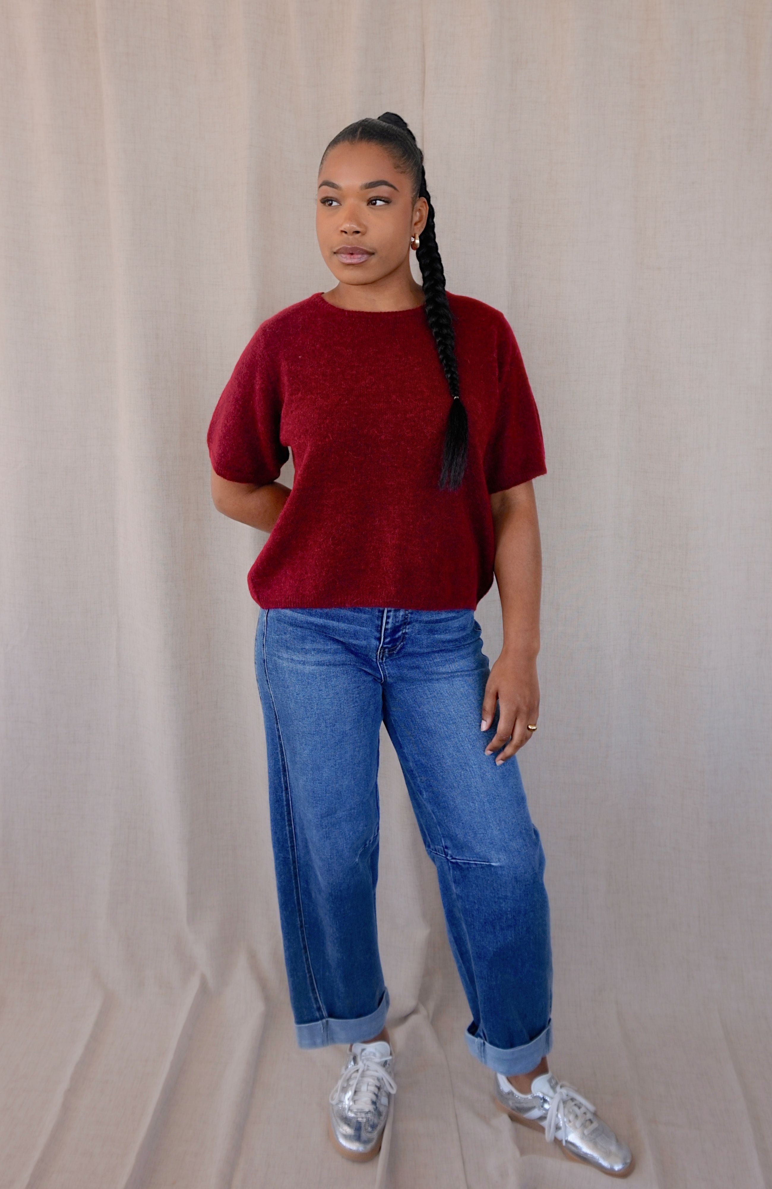 A woman modeling a short-sleeve crewneck knit sweater in deep burgundy, paired with blue jeans and silver sneakers, standing against a neutral beige fabric backdrop.