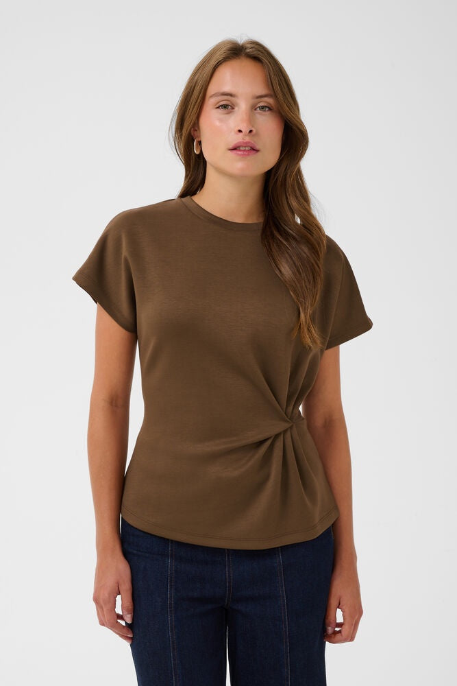 A woman modeling a short-sleeve brown knit top with a twisted detail at the waist, paired with dark blue jeans, against a light gray studio background.