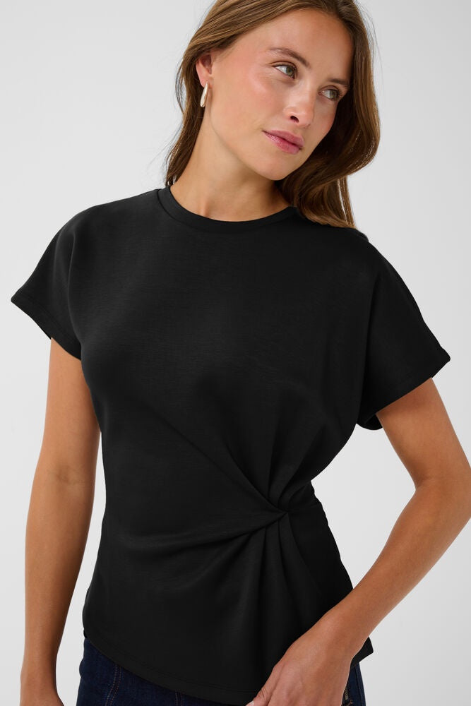 A woman wearing a black short-sleeve crewneck top with a twisted knot detail at the waist, photographed against a light gray studio background.