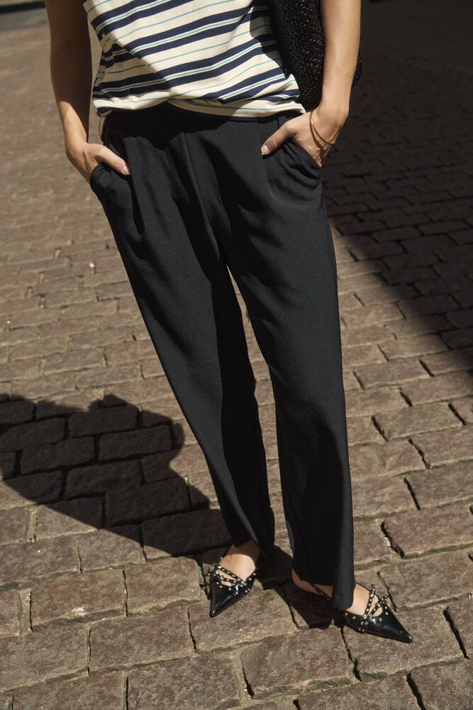 A person wearing black wide-leg trousers with a striped top and pointed flats, standing on a cobblestone street in bright sunlight.