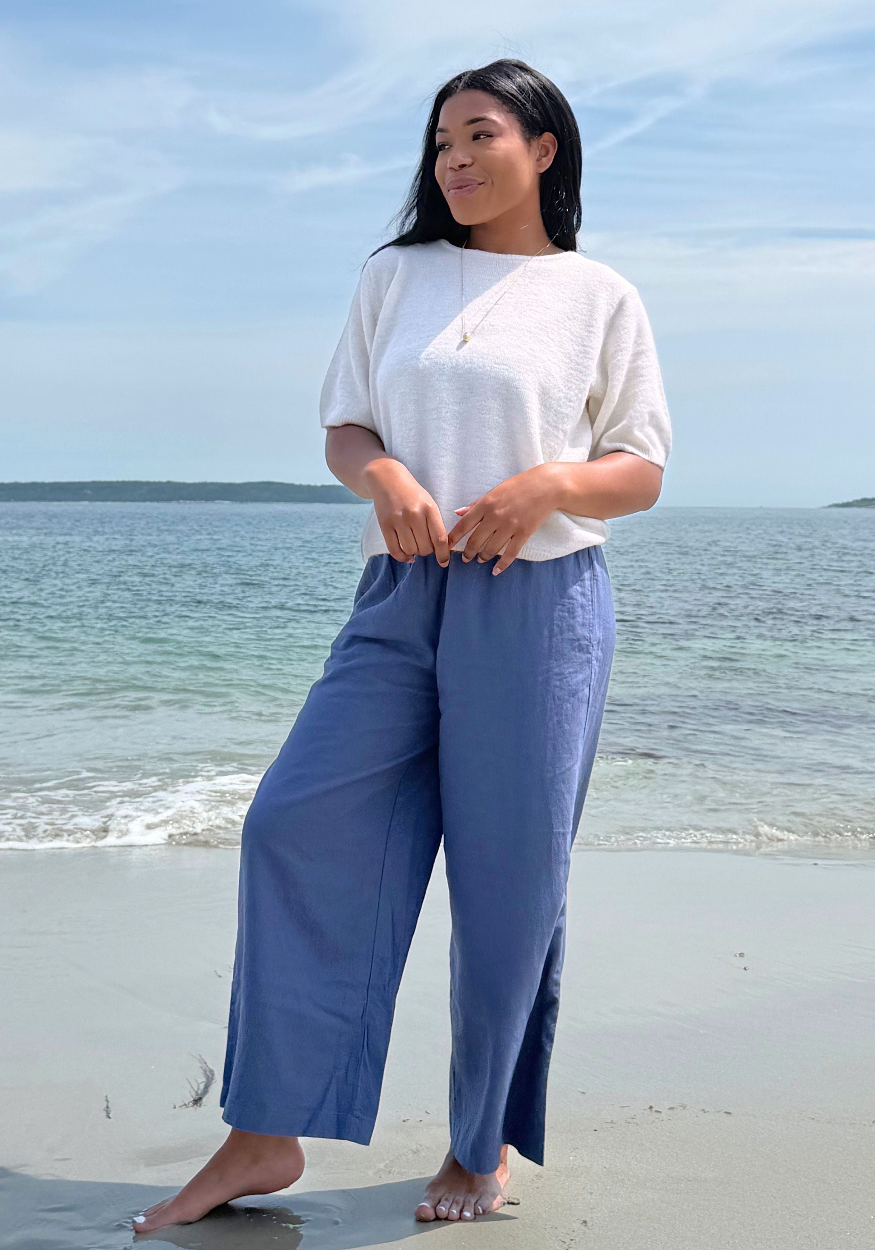A woman stands barefoot on a sandy beach wearing a white short-sleeve top and blue wide-leg pants.