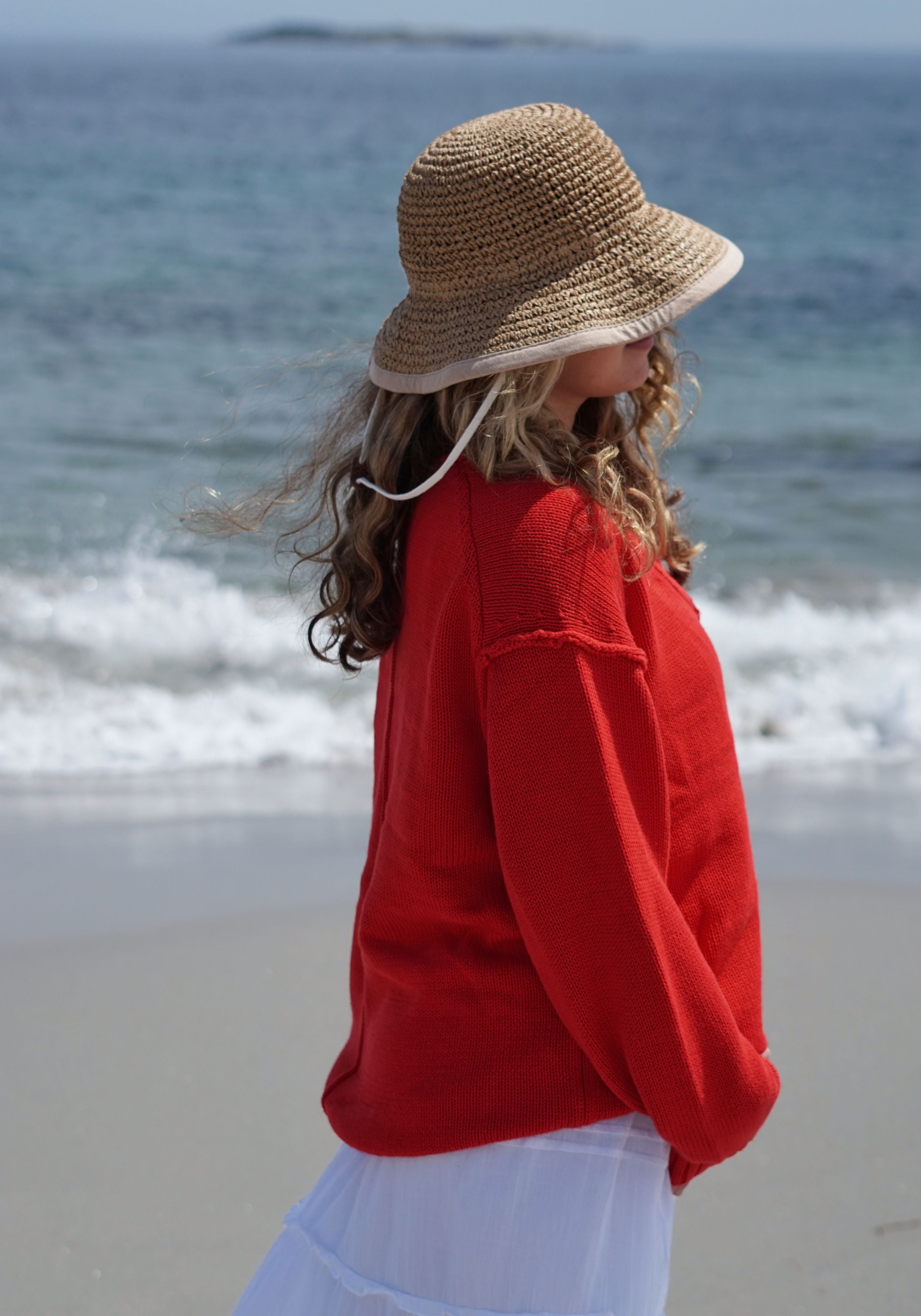 A woman standing on a beach wearing a woven straw sun hat, a bright red knit sweater, and a white skirt.