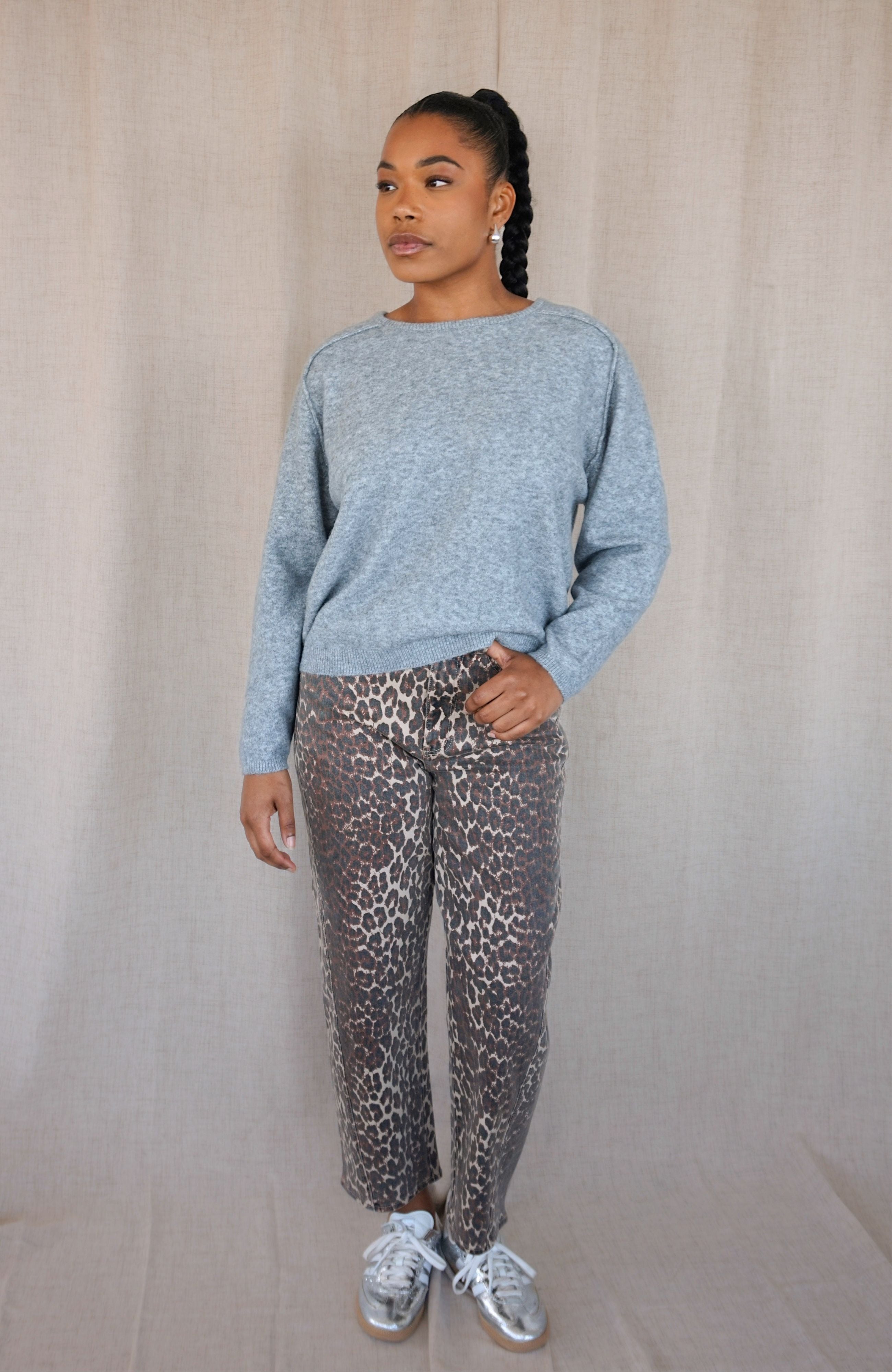 A woman modeling a light grey heathered knit sweater and brown leopard-print pants with silver sneakers, standing against a neutral beige fabric backdrop.