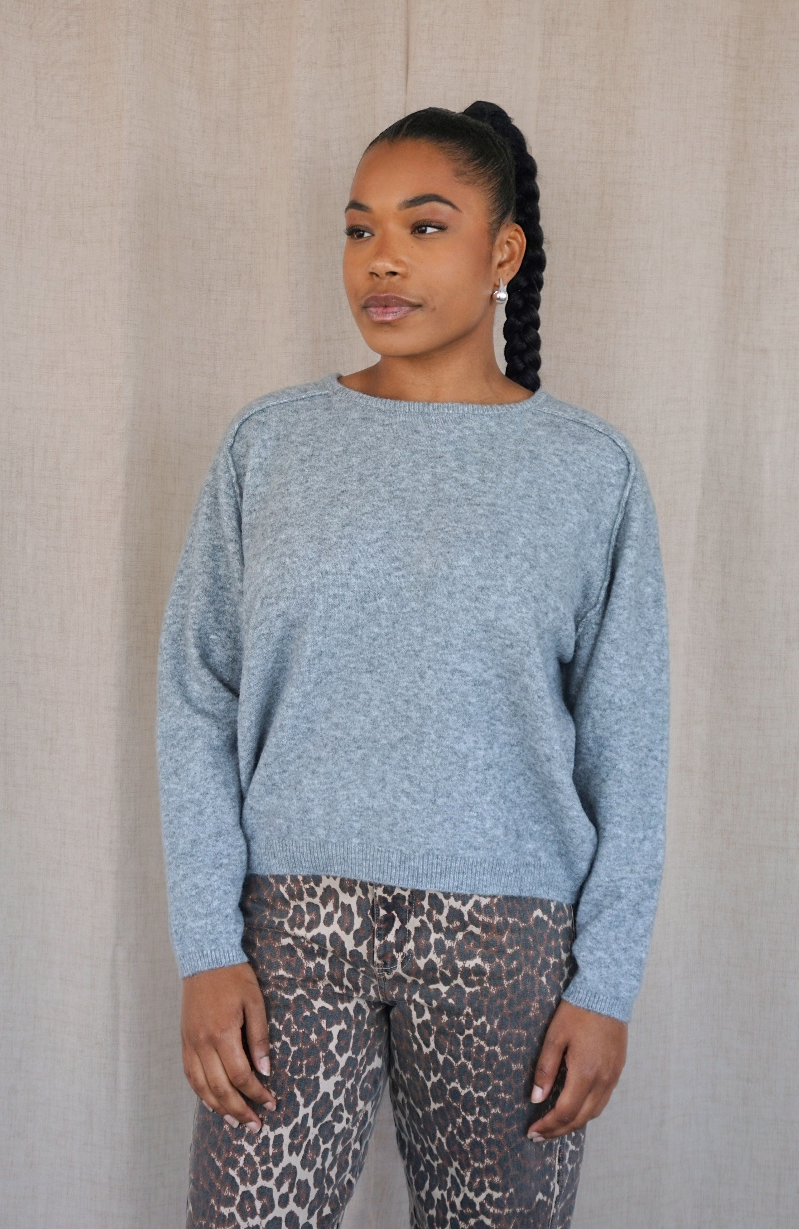 A woman wearing a light grey heathered knit sweater and leopard print pants, standing against a neutral beige textured backdrop.