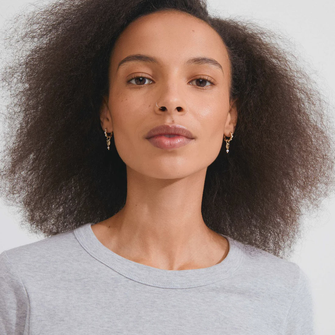 A close-up portrait of a woman with voluminous dark curly hair wearing small gold hoop earrings and a light gray crewneck top against a plain light gray background.