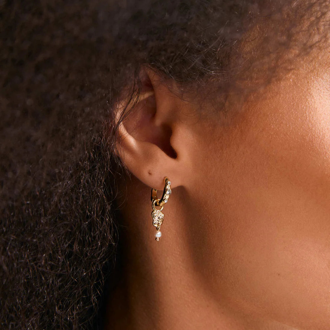 A close-up of a person's ear wearing a small gold huggie hoop earring adorned with clear stones and a dangling pearl.