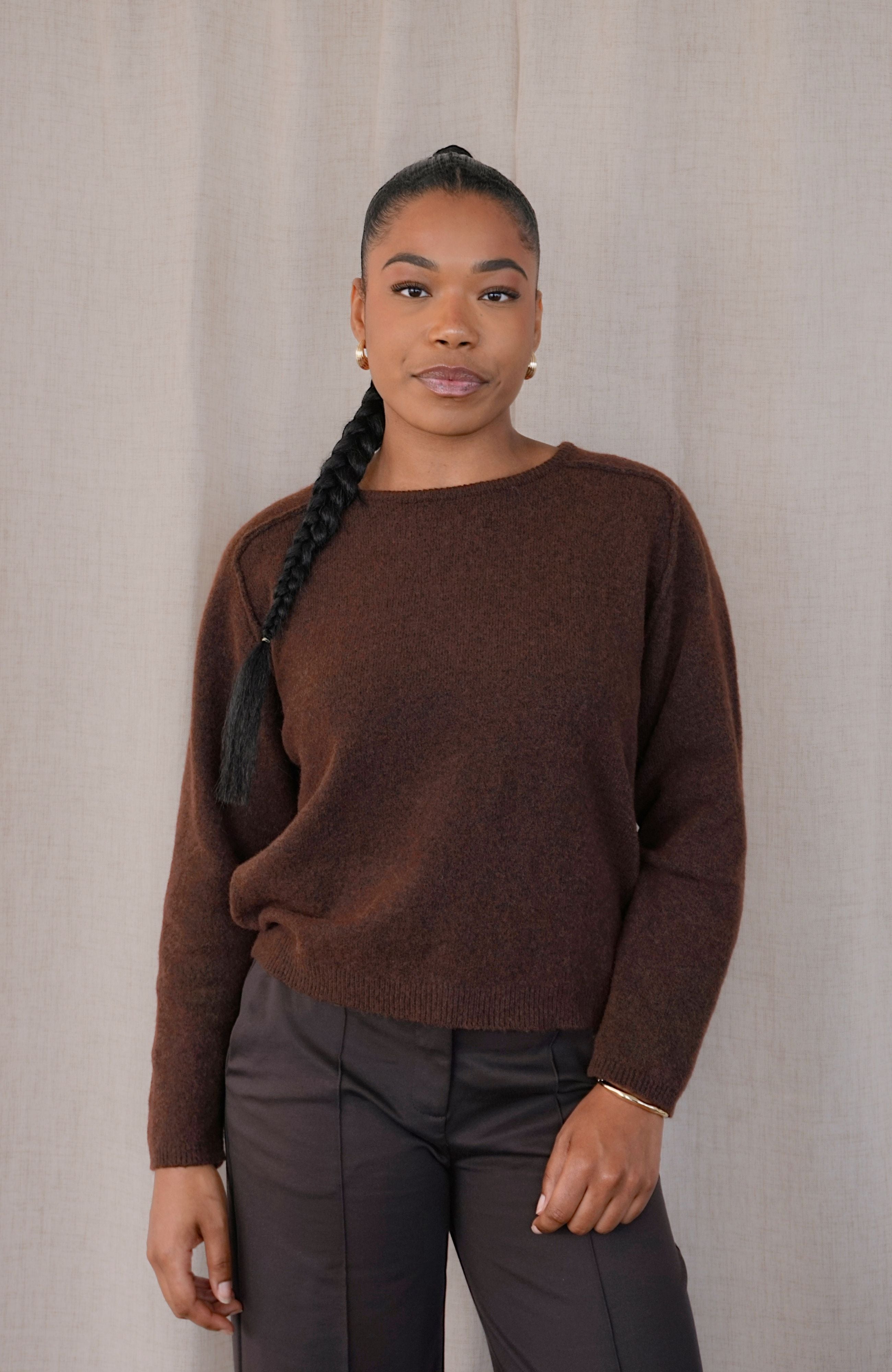 A woman wearing a brown knit crewneck sweater and dark gray trousers, standing against a neutral beige fabric backdrop.