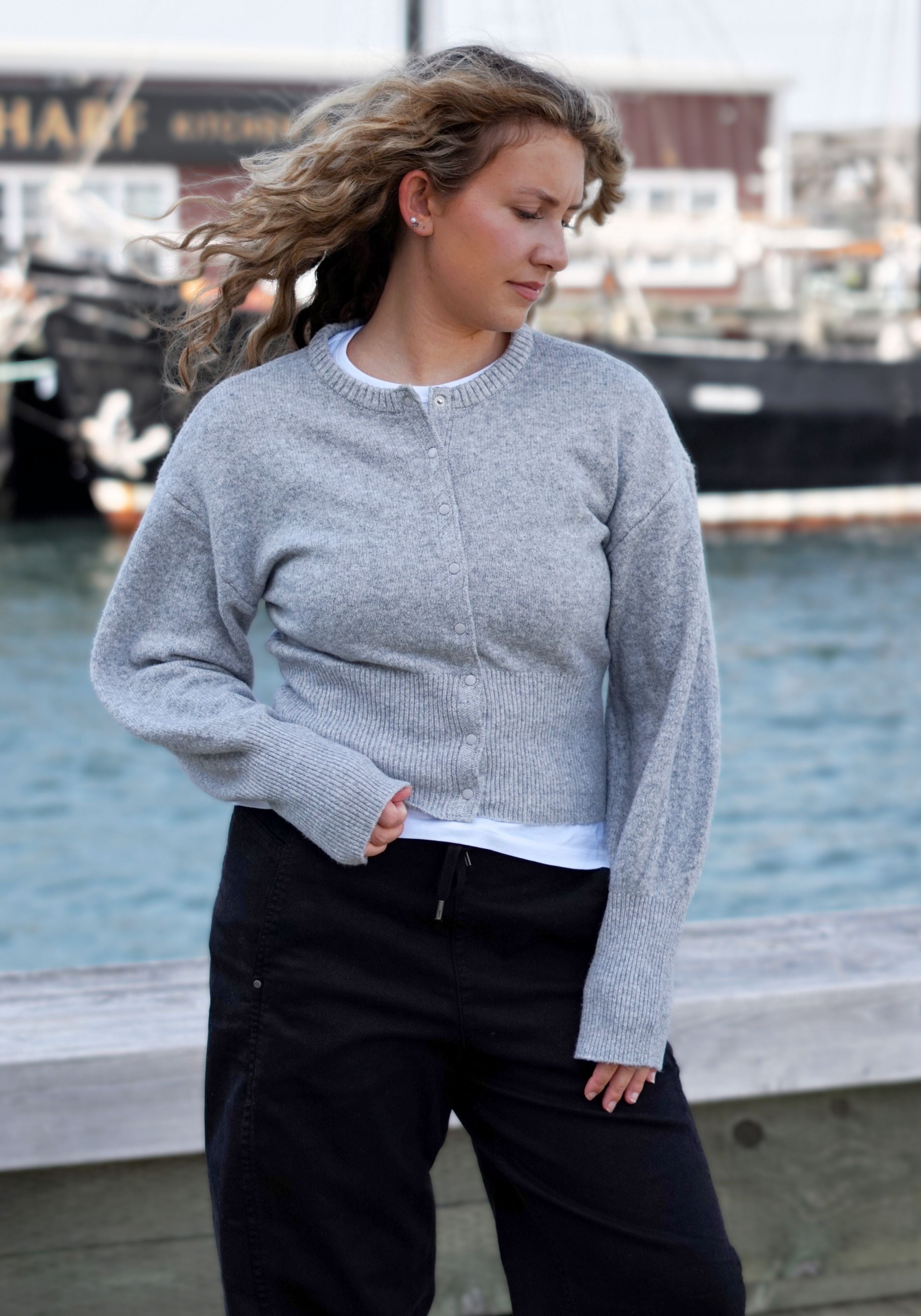 A woman wearing a light gray knit cardigan sweater and dark pants, standing outdoors.