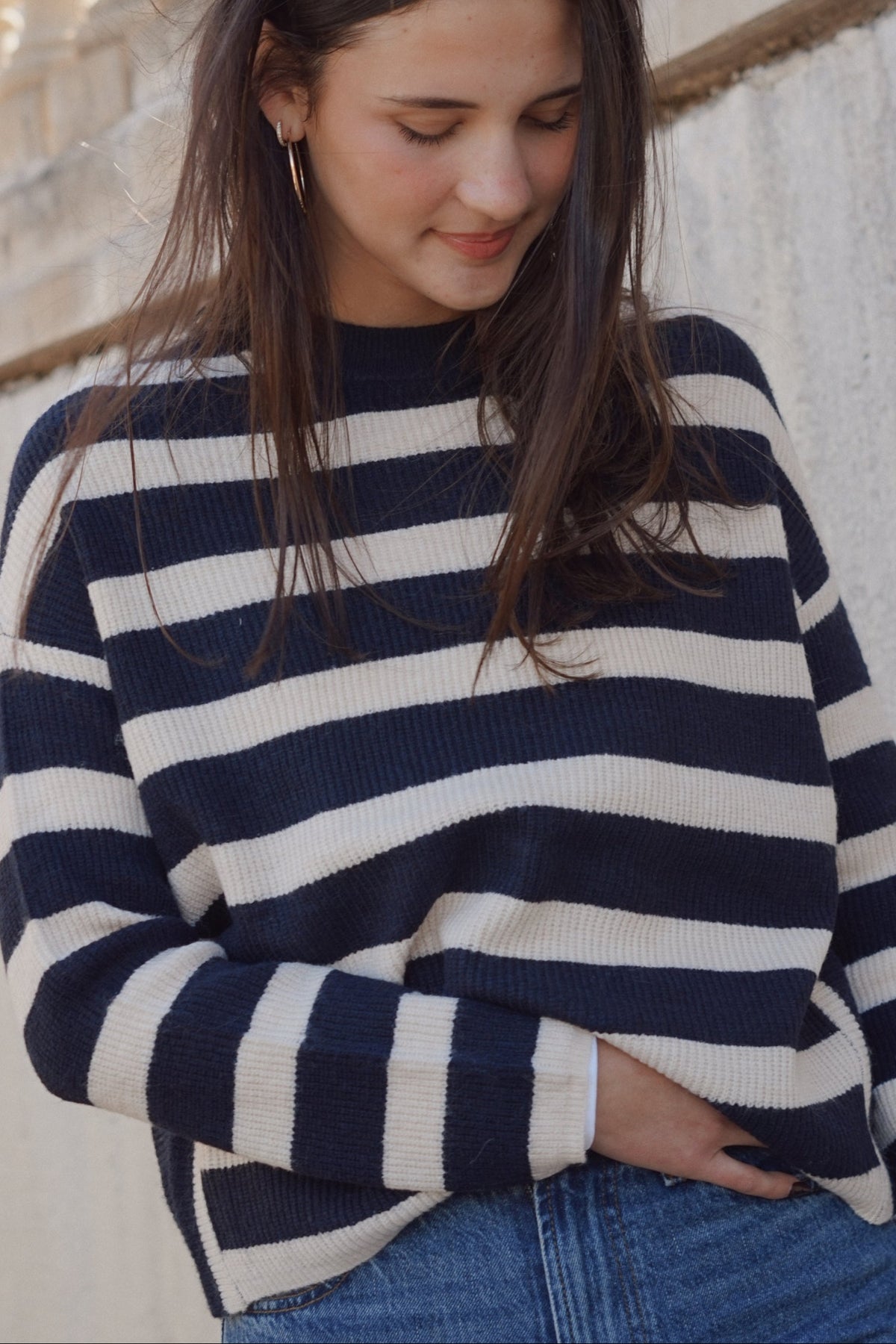 A woman wearing a navy blue and off-white horizontally striped knit sweater, paired with blue jeans, standing against a light-colored stone wall.