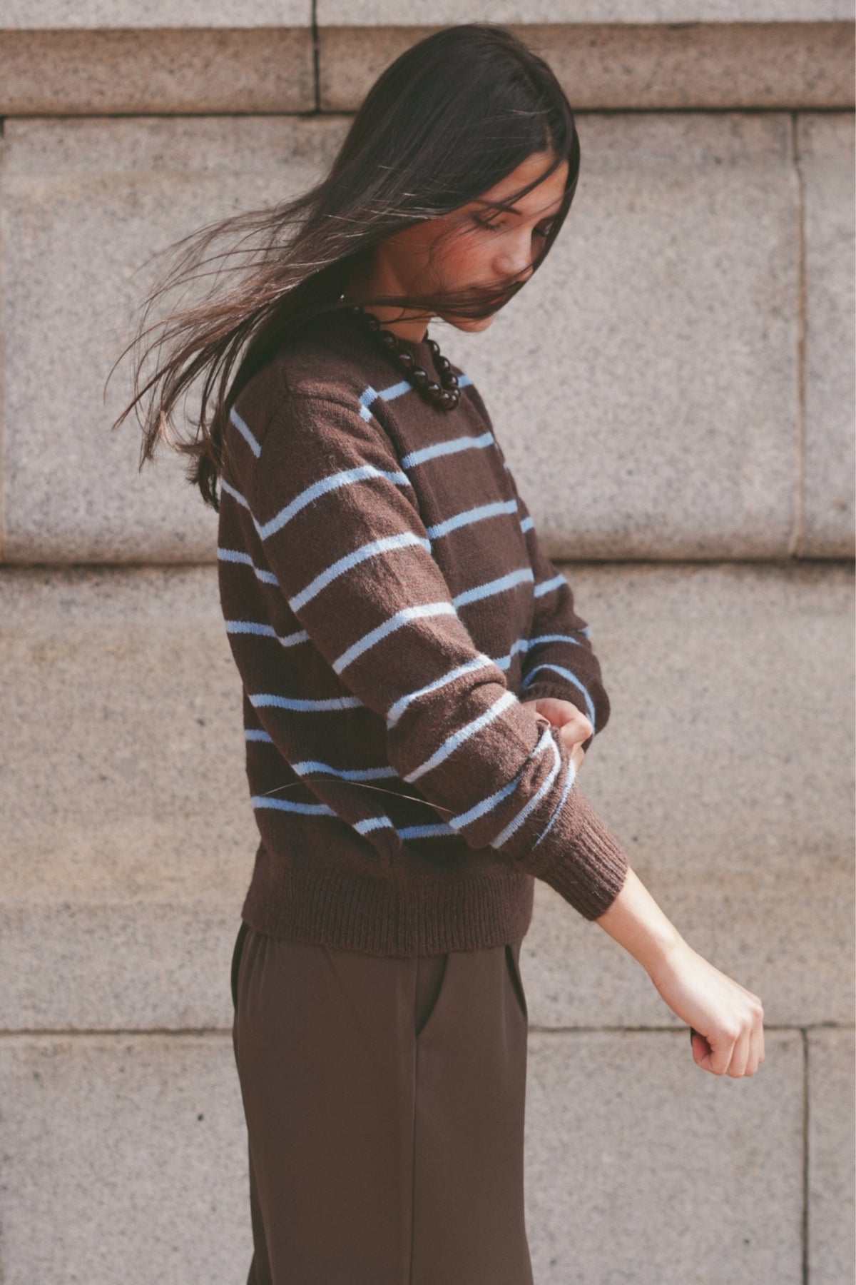 A woman wearing a brown knit sweater with light blue horizontal stripes and dark brown trousers.