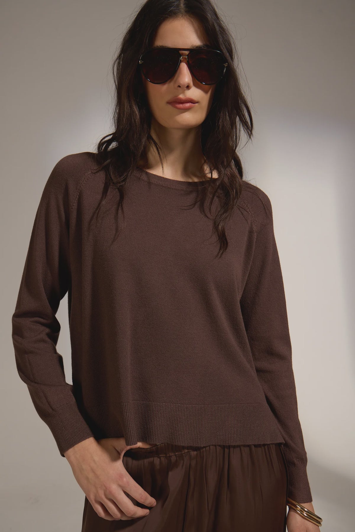 Woman wearing a brown long-sleeve top and sunglasses against a neutral background