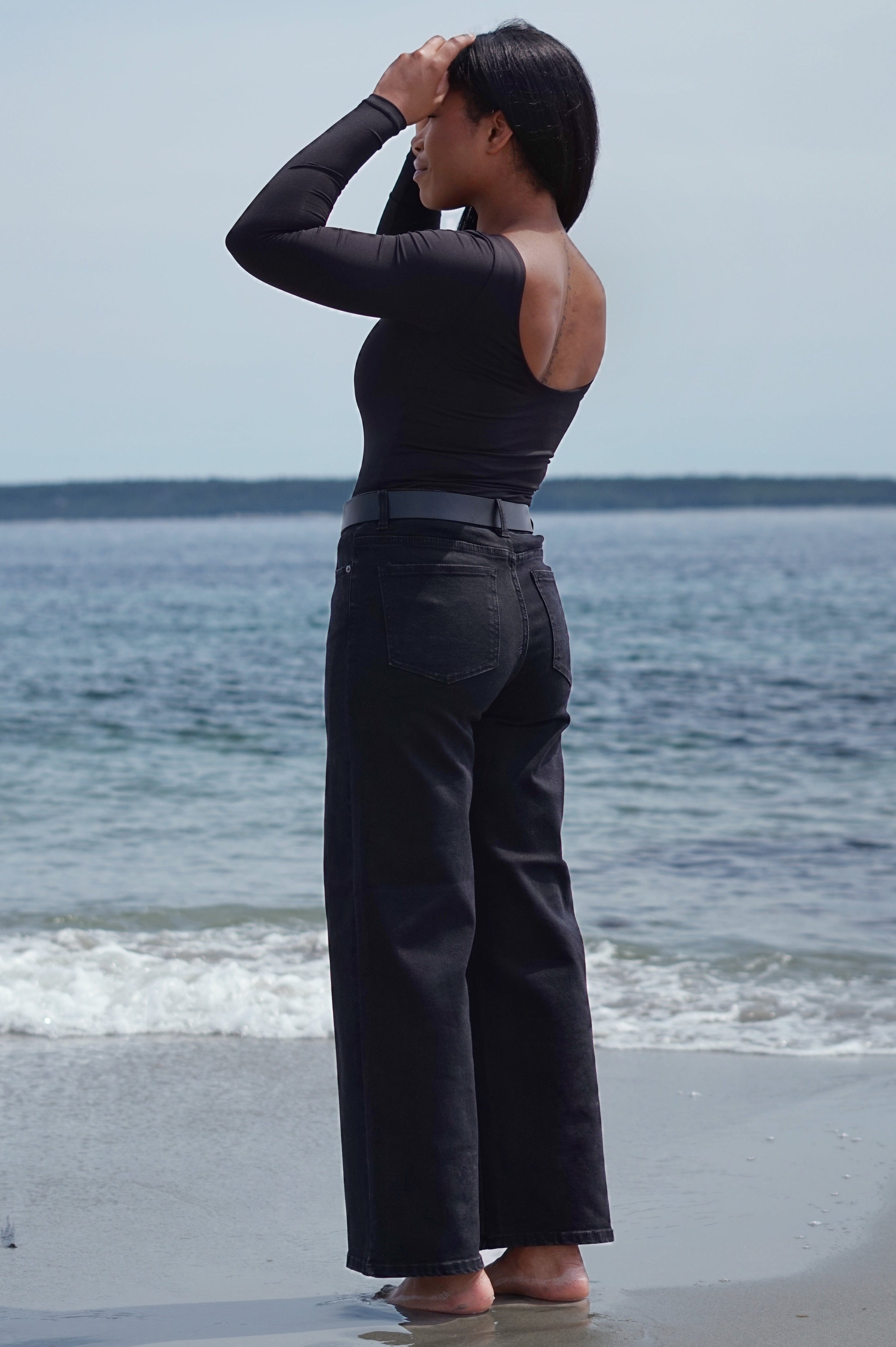 A woman stands on a beach facing the ocean, wearing a black long-sleeve top with cutout details and black wide-leg jeans.