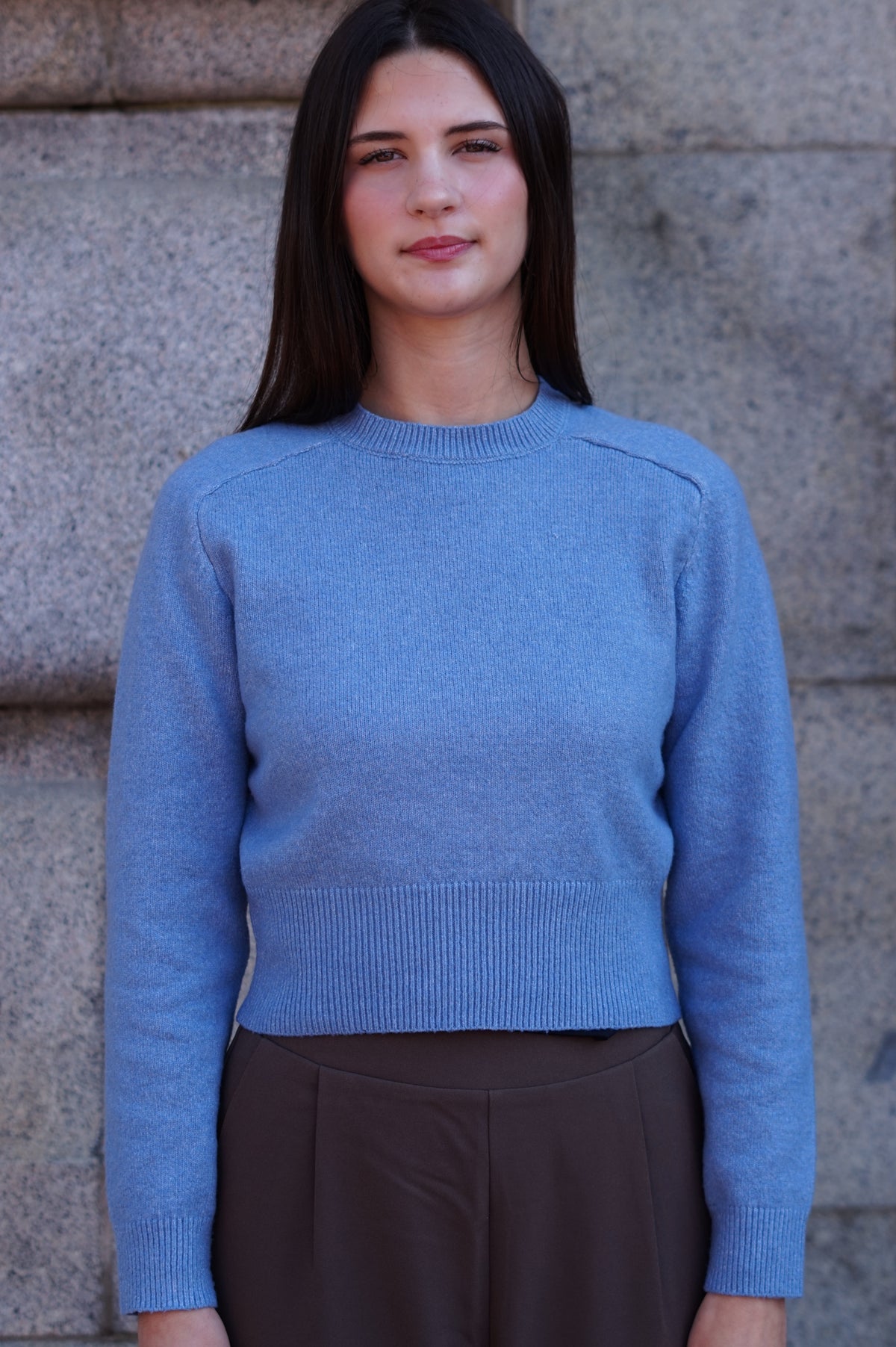 A woman with long dark hair wearing a light blue ribbed knit sweater and dark brown trousers, standing against a gray stone wall.
