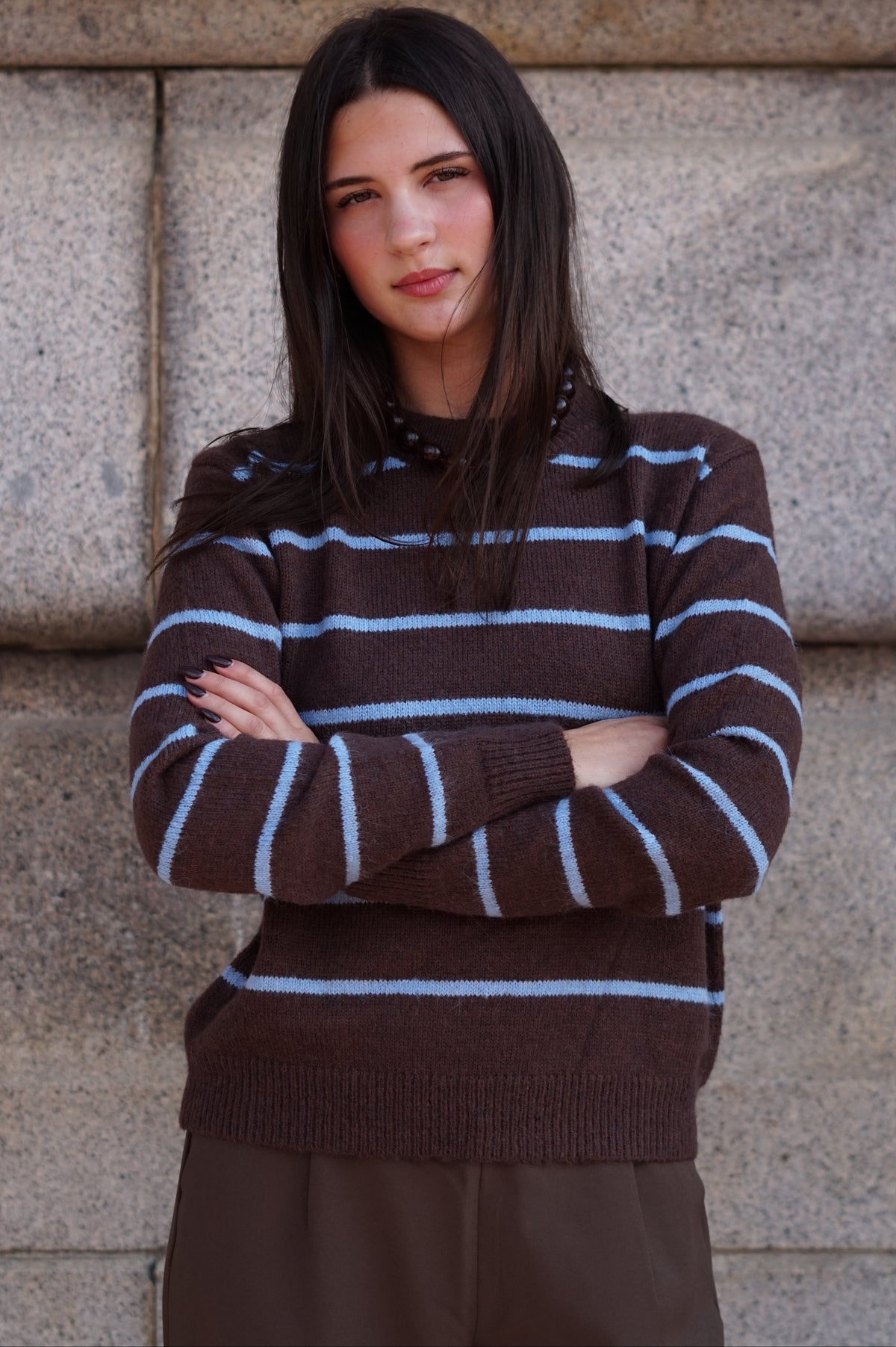 A woman wearing a brown knit sweater with light blue horizontal stripes and dark brown pants.